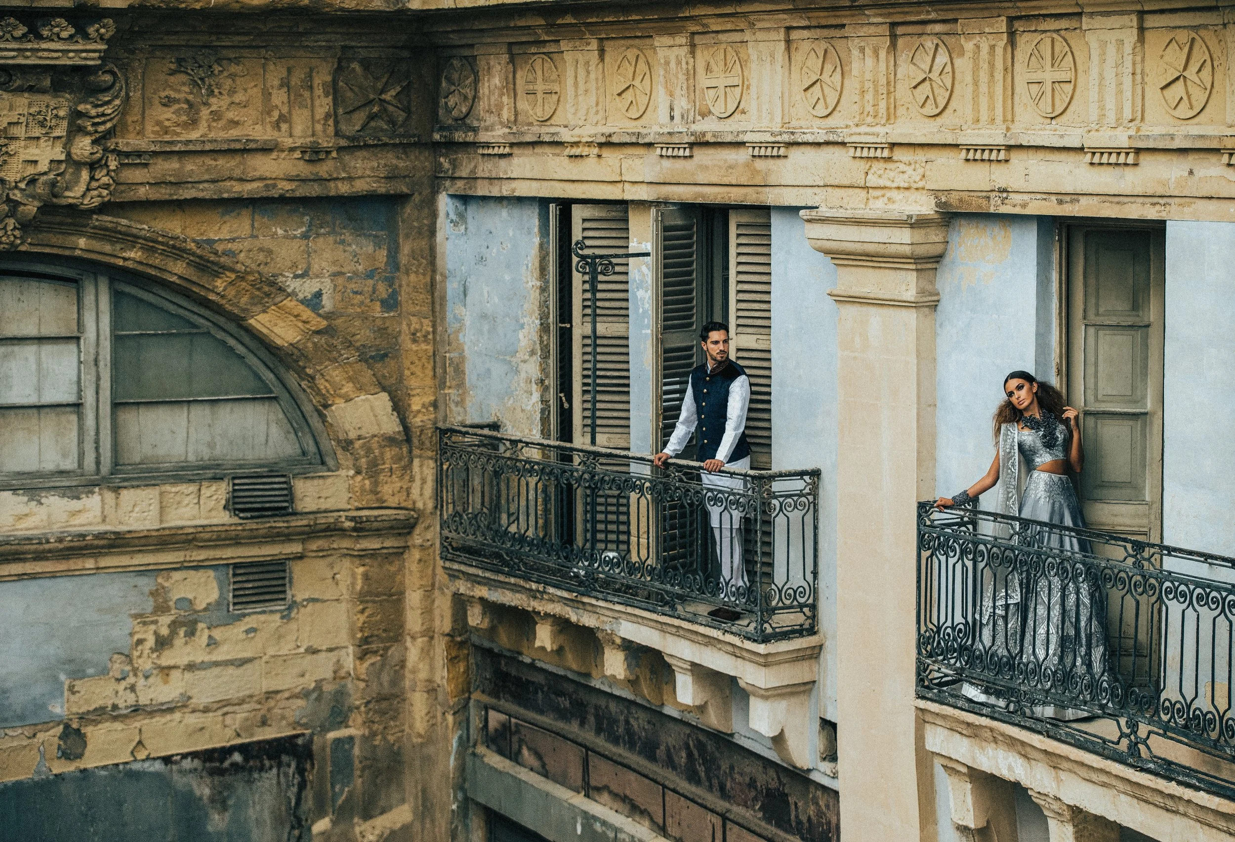 A man and woman in formal attire standing on a vintage balcony of an old, rustic building with weathered walls and ornate architectural details.