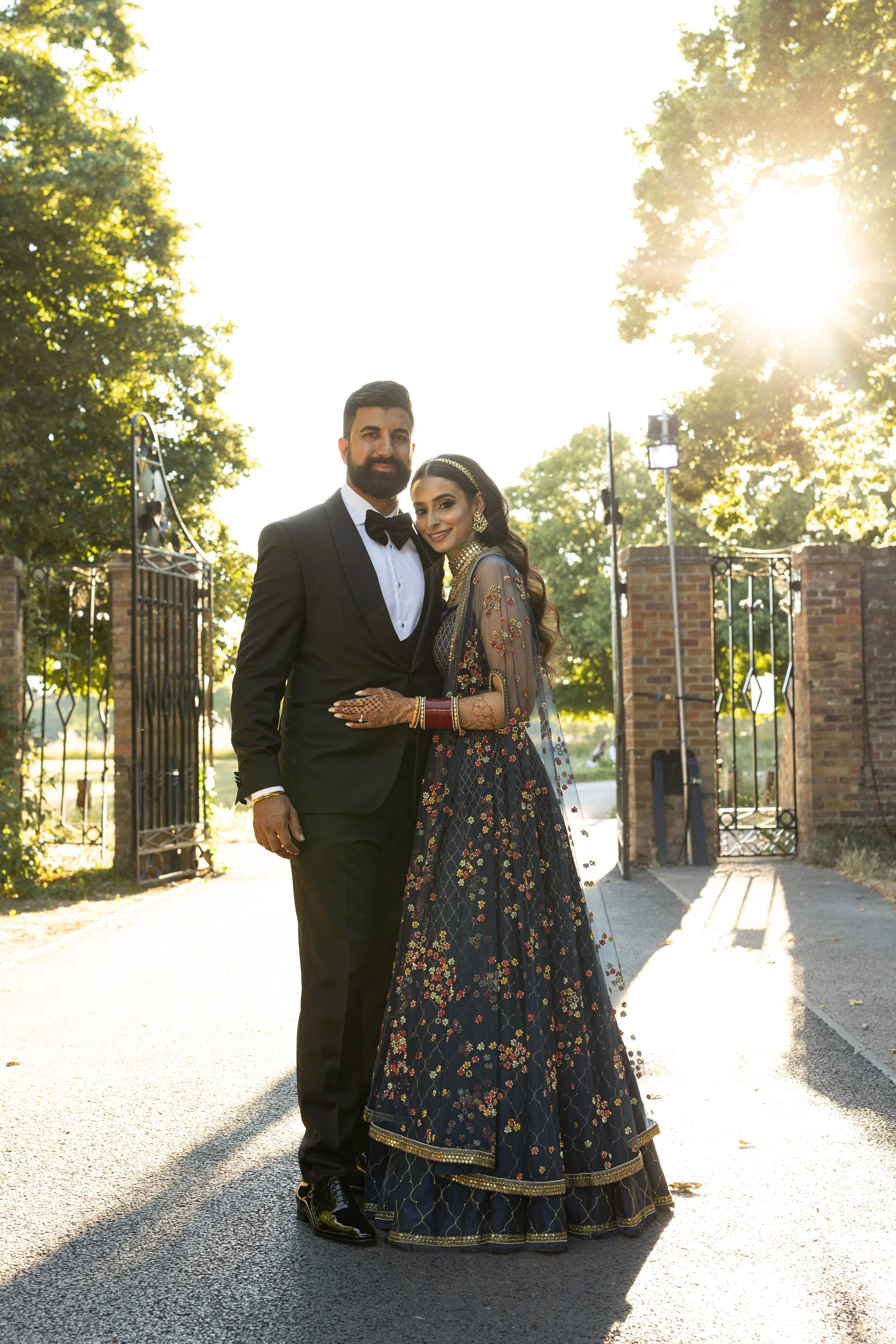 A well-dressed couple, in formal attire, standing in front of an open gate with sunlight shining through the trees behind them.