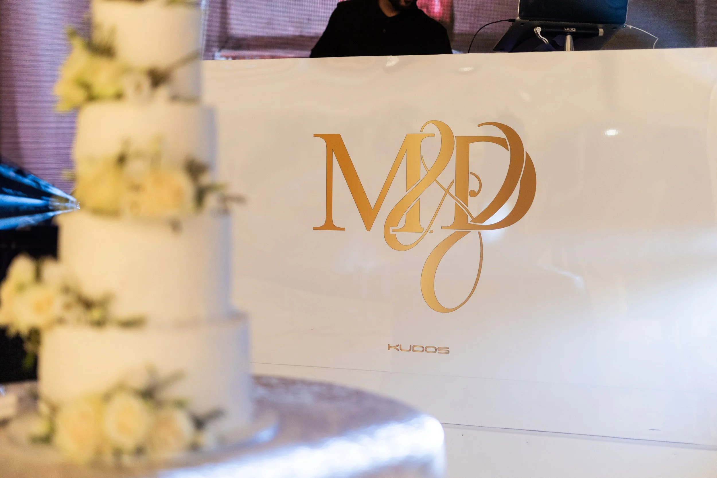 A white cake with yellow and white flowers in the foreground on the left side, and a white sign with gold lettering reading 'MD' and 'KUDOS' in the background. The cake is slightly out of focus, while the sign is clear and centered.
