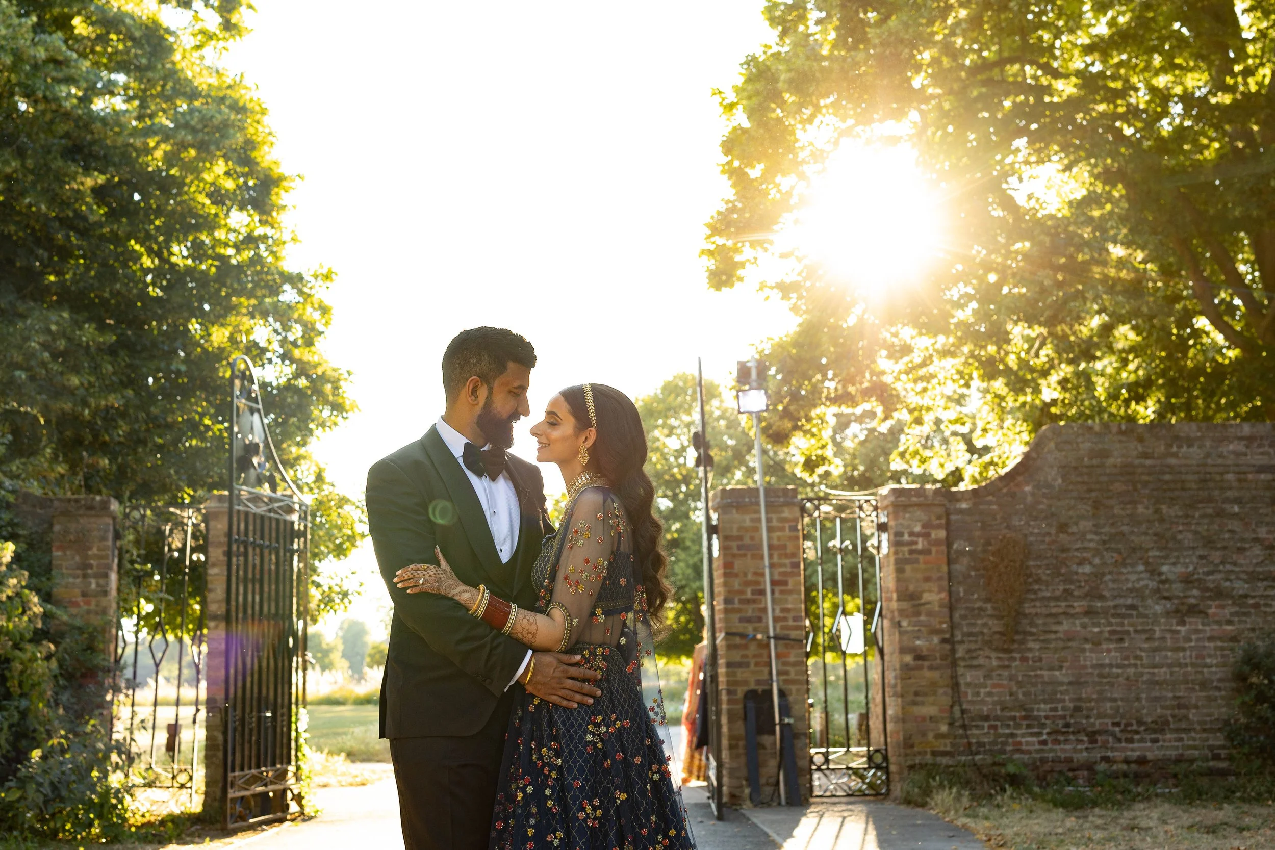 A bride and groom in wedding attire standing close together outdoors with sunlight shining through trees behind them.