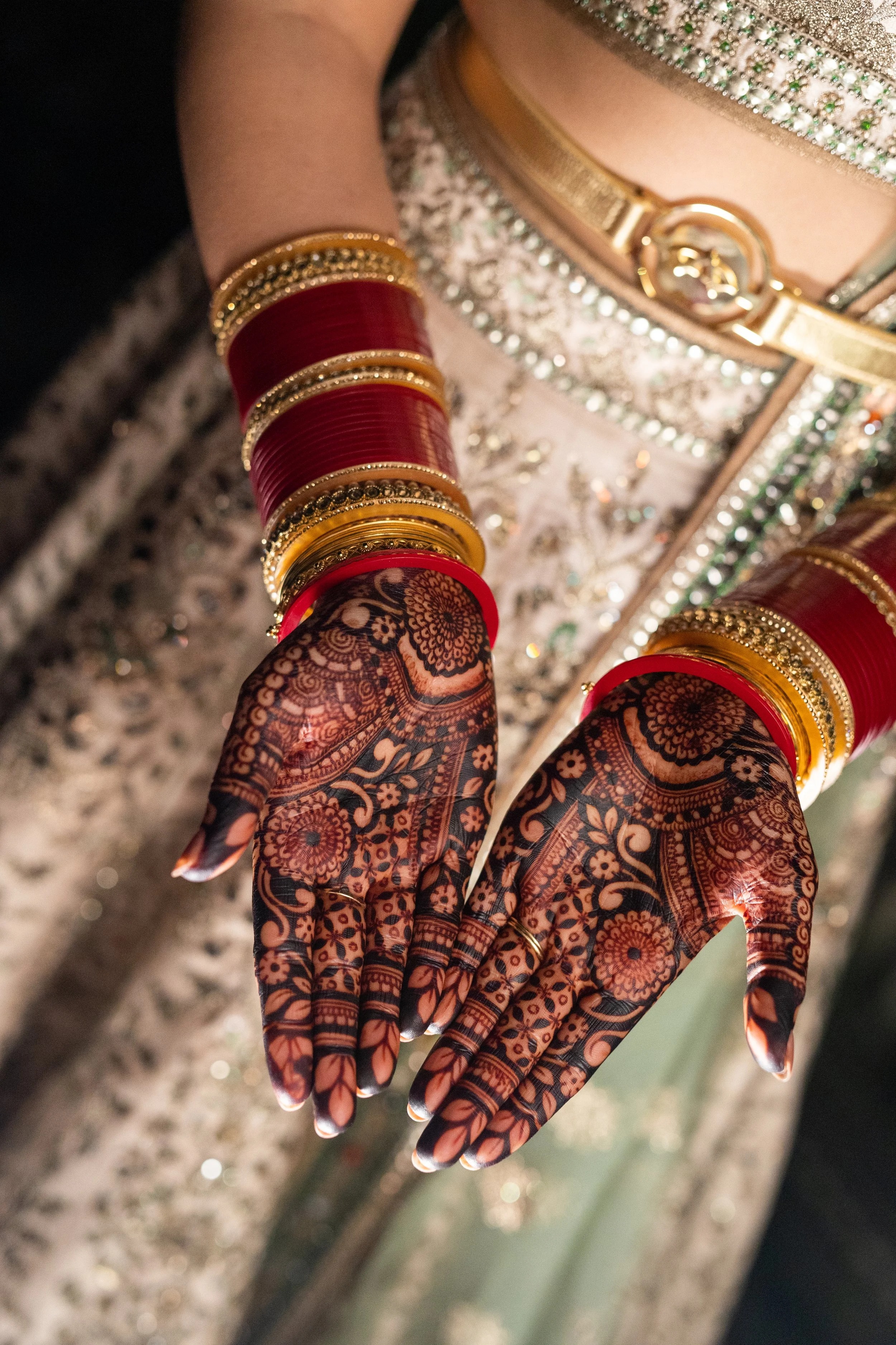 Close-up of hands decorated with intricate mehndi (henna) designs, wearing multiple red and gold bangles, against a background of traditional attire with embellishments.