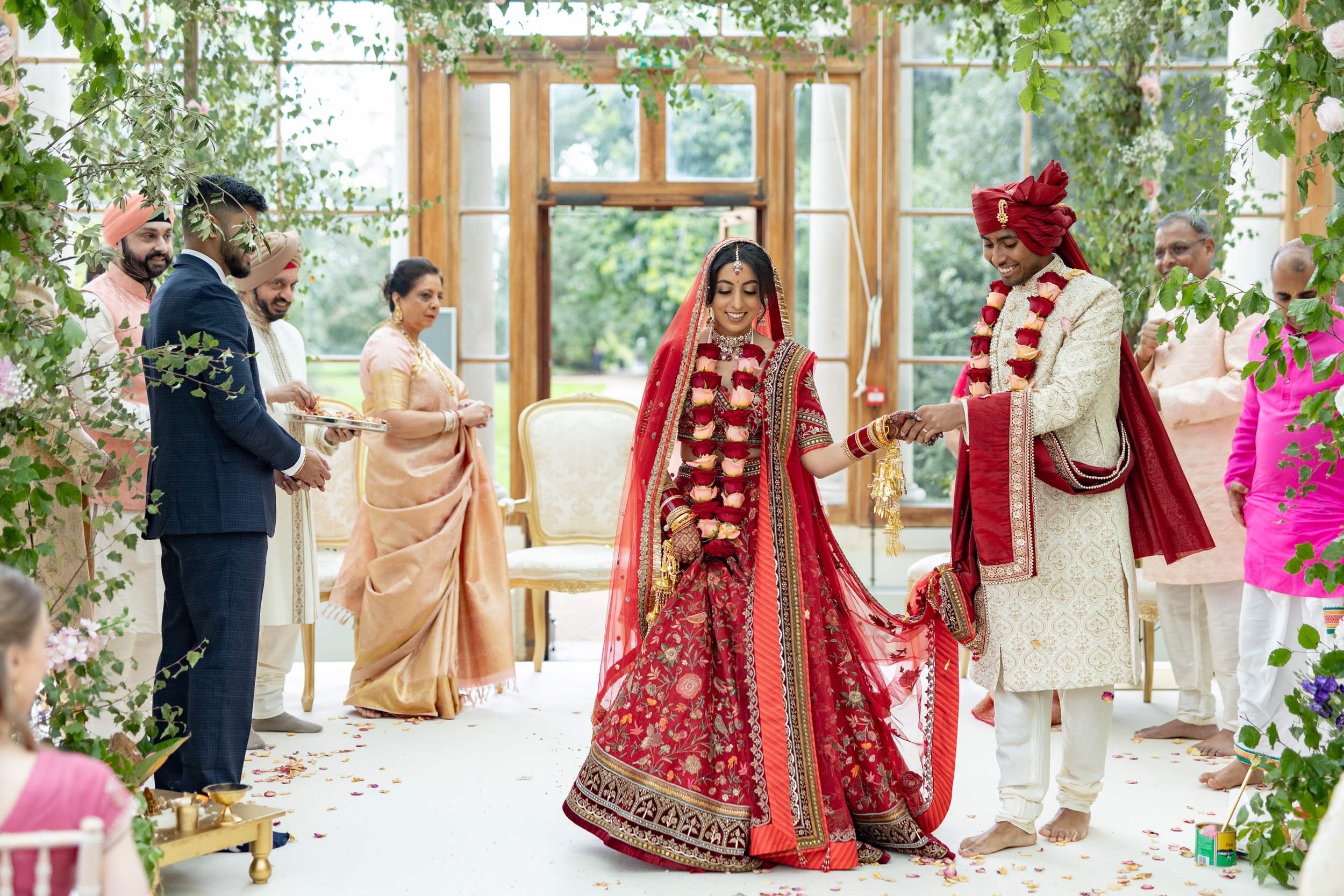 Indian wedding ceremony with bride and groom exchanging rings, surrounded by family and friends in traditional attire, indoors decorated with greenery and flowers.