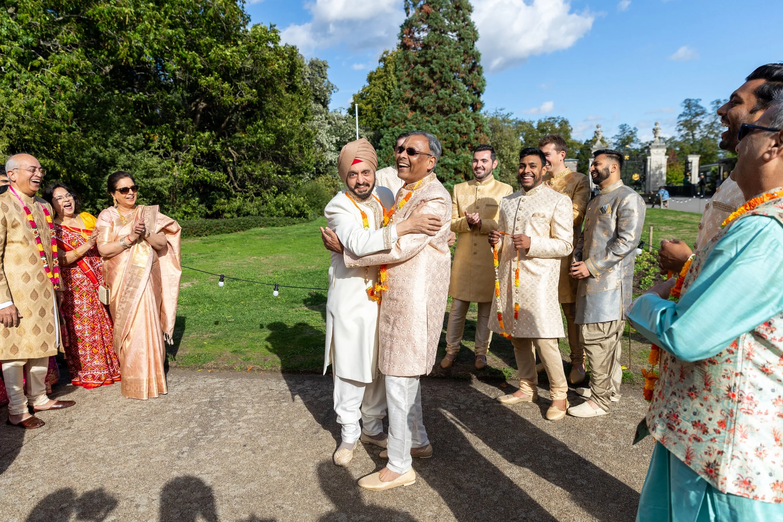 Group of people in traditional Indian attire celebrating outdoors, with two men hugging in the center, surrounded by others clapping and smiling.