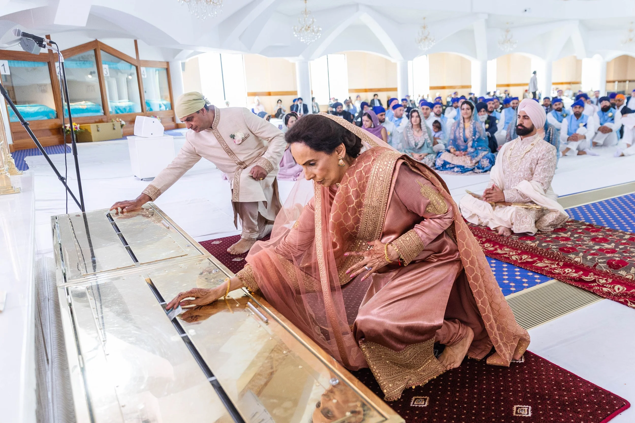 A traditional Indian wedding ceremony with the bride, groom, and family members in ceremonial attire, seated on carpets, participating in rituals inside a decorated hall with seated guests in the background.