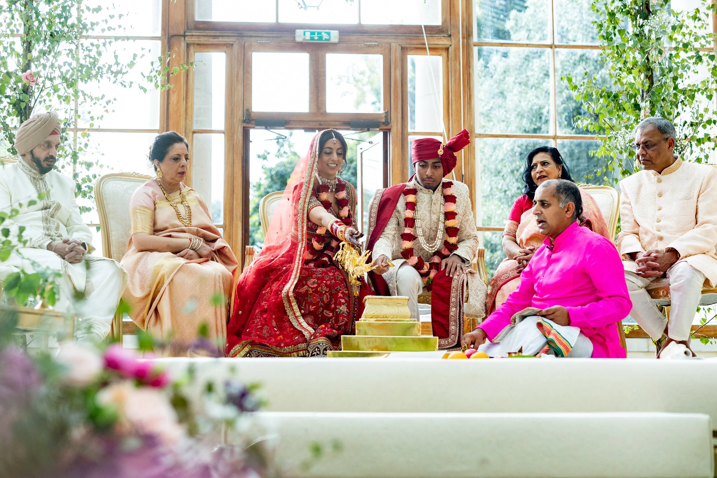 Indian wedding ceremony with couple in traditional attire sitting on a stage, surrounded by family members, inside a bright room with large windows and greenery outside.