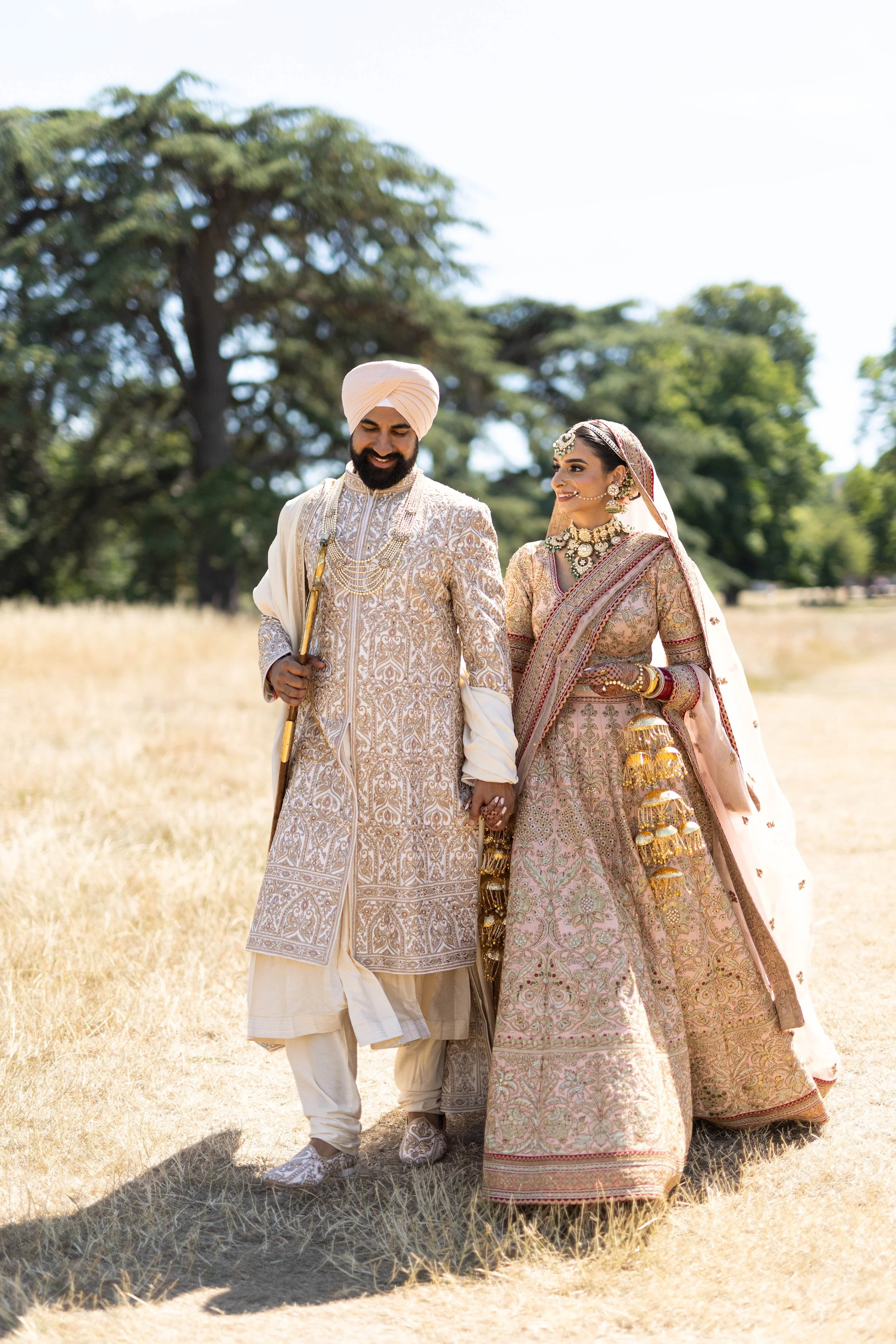 A smiling South Asian couple dressed in traditional wedding attire, holding hands and walking in an outdoor grassy field with large trees in the background.