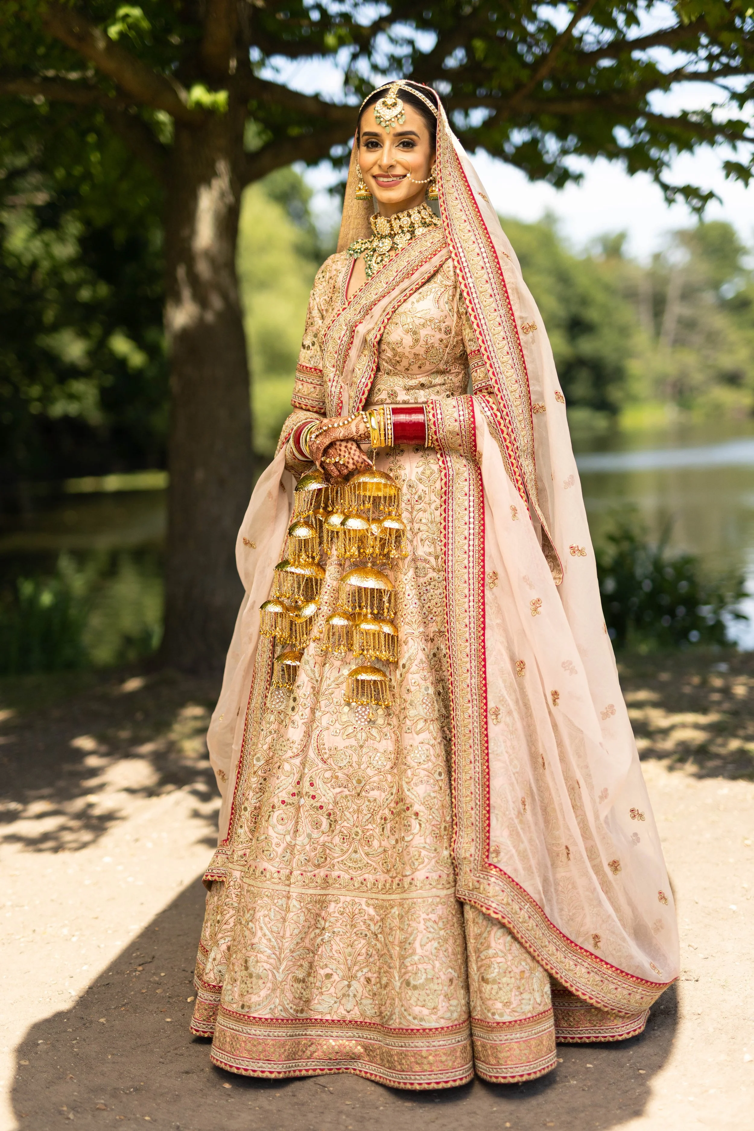 A woman dressed in traditional Indian bridal attire, wearing a heavily embroidered cream and gold bridal lehenga, with jewelry including a necklace, earrings, nose ring, and bangles, standing outdoors near a tree and a body of water.