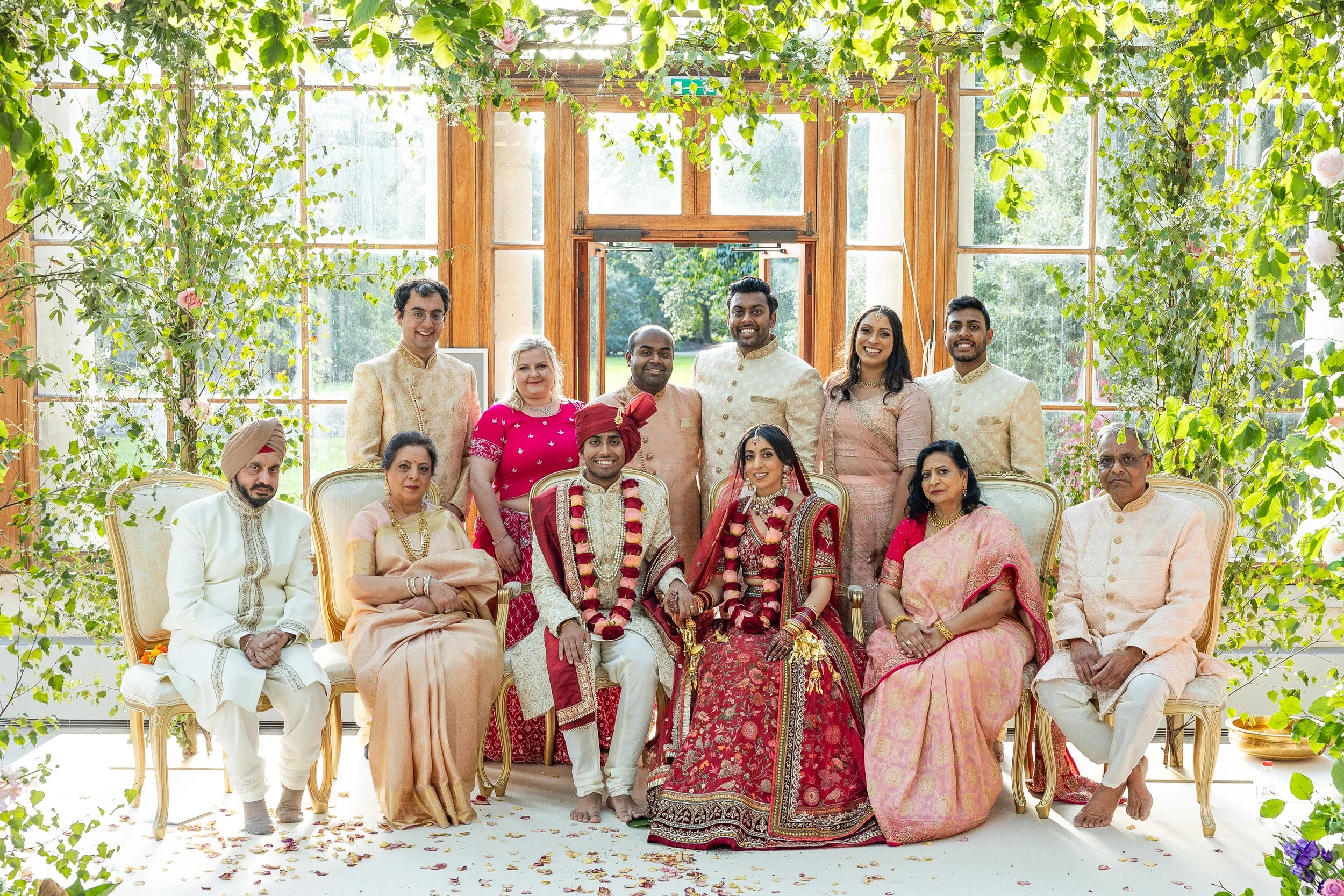 Indian wedding family group with bride and groom seated in center, surrounded by relatives on a bright, flower-decorated porch with green foliage and large windows in the background.
