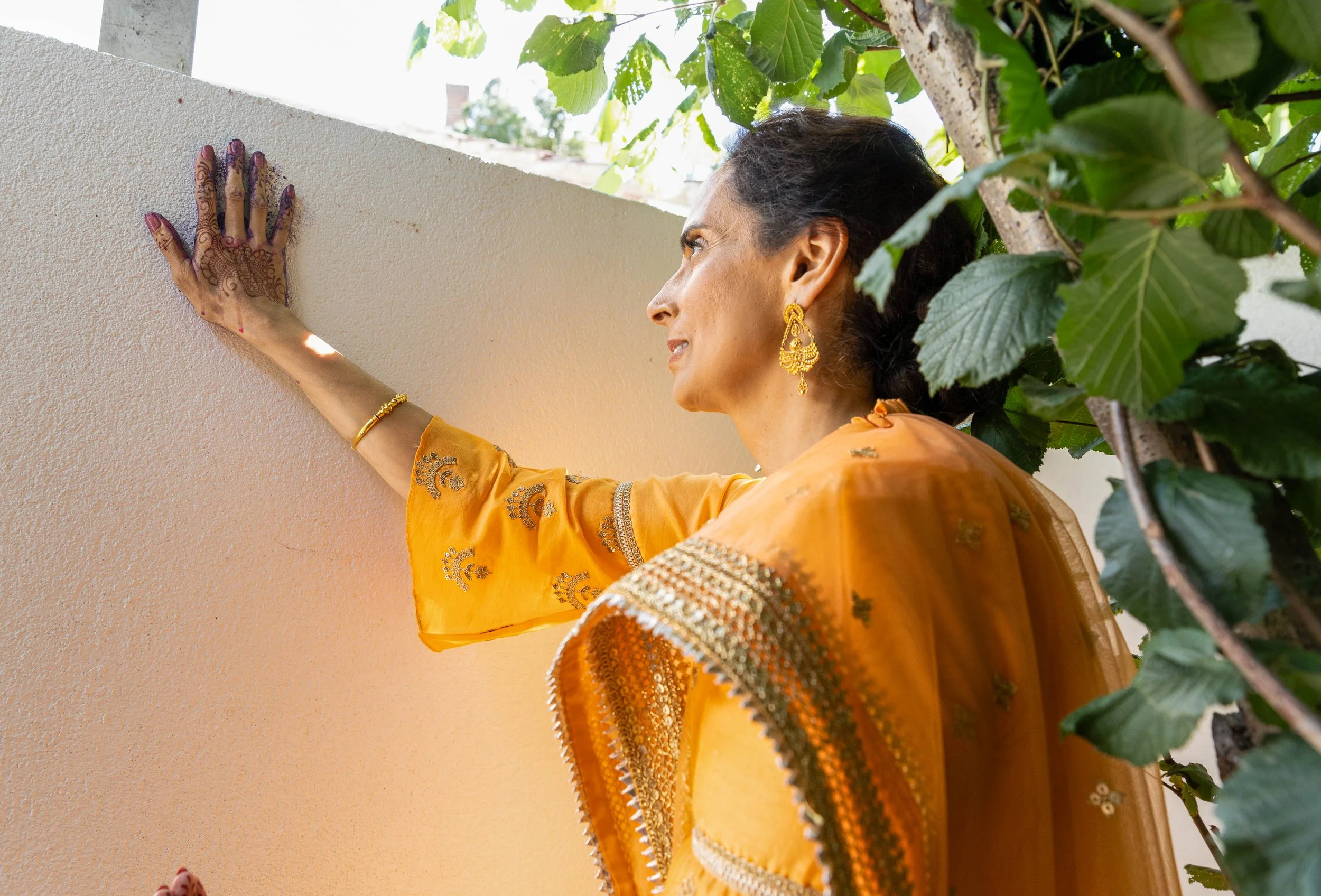 A woman wearing a yellow traditional dress with jewelry, including earrings and bangles, reaching up to touch a wall with her hand. She is standing near some green foliage.