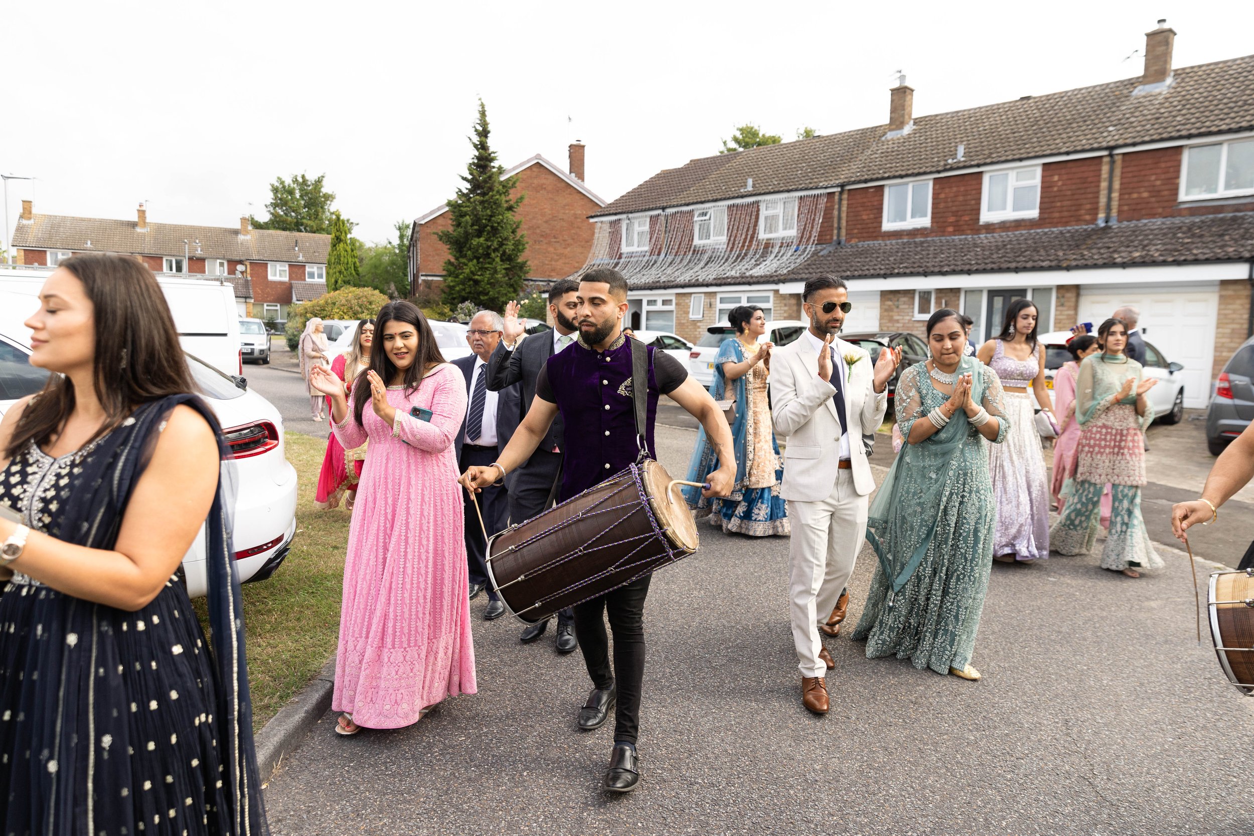 A group of people dressed in traditional Indian attire, participating in a wedding procession with drummers, walking on a residential street.