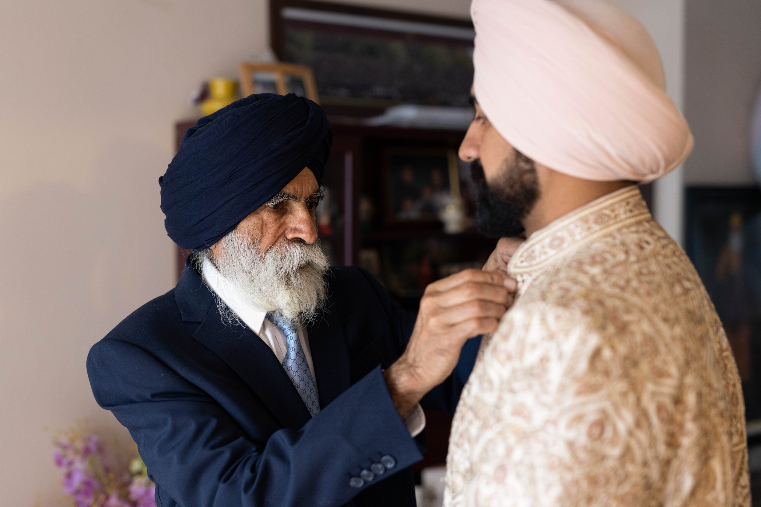 An elderly man of Sikh heritage wearing a navy blue turban and suit, adjusting the collar of a younger man of Sikh heritage wearing a light pink turban and traditional attire, indoors in a warmly lit room.