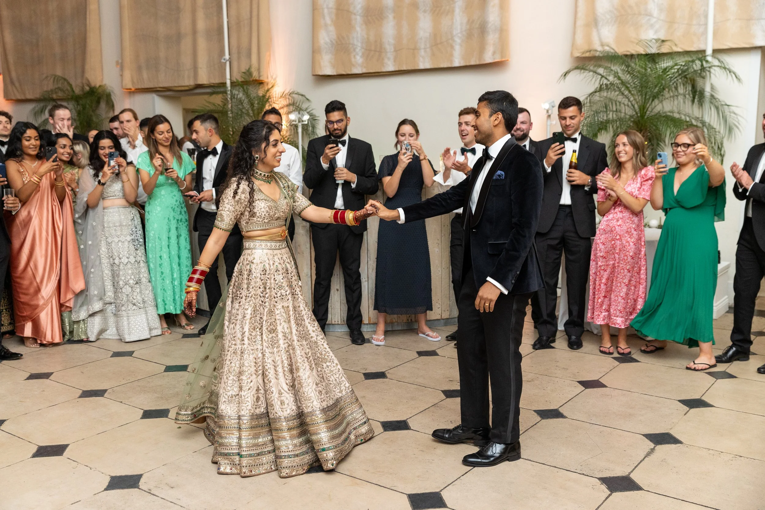 A couple dancing at a wedding reception, with guests watching and taking photos in the background.