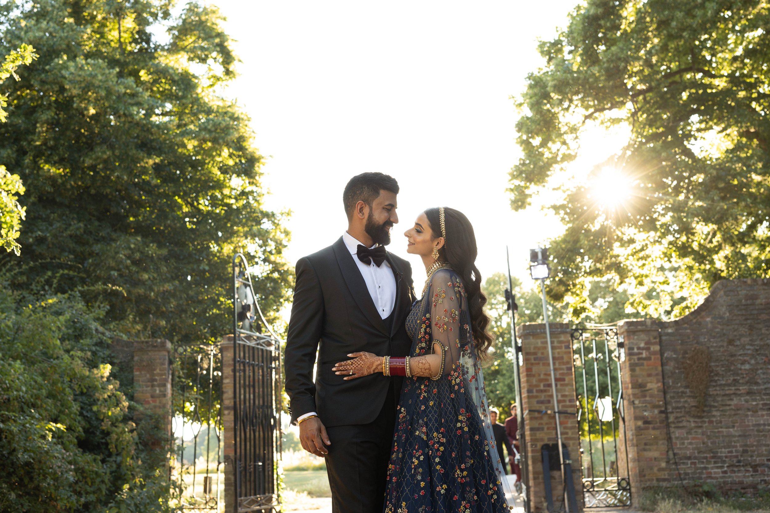 A couple dressed in formal attire, a man in a suit and a woman in traditional Indian clothing, standing closely and smiling at each other outdoors with sunlight filtering through trees in the background.
