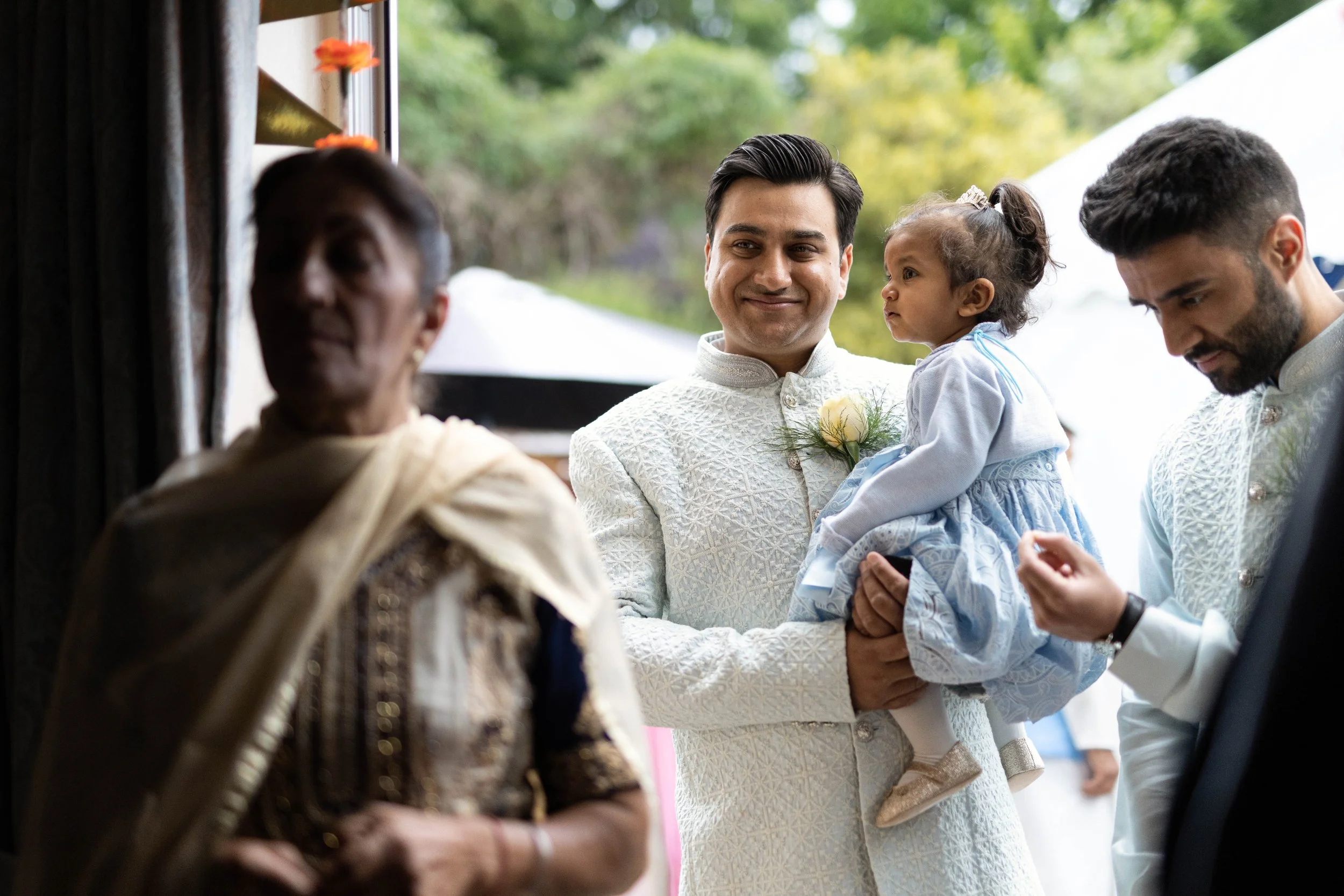 A man in traditional Indian attire holding a young girl, also in traditional attire, while others around them are engaged in conversation; outdoors with greenery in the background.