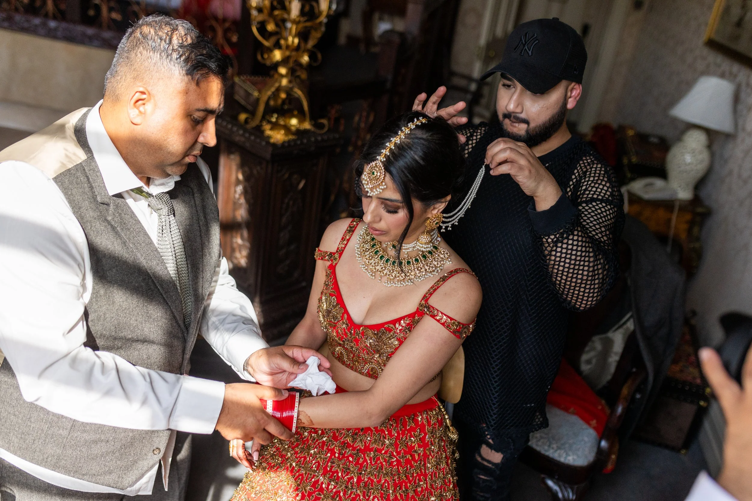 A bride dressed in traditional red and gold jewelry and attire, receiving help from a man in a gray vest and white shirt, with another man adjusting her veil in a decorated indoor setting.