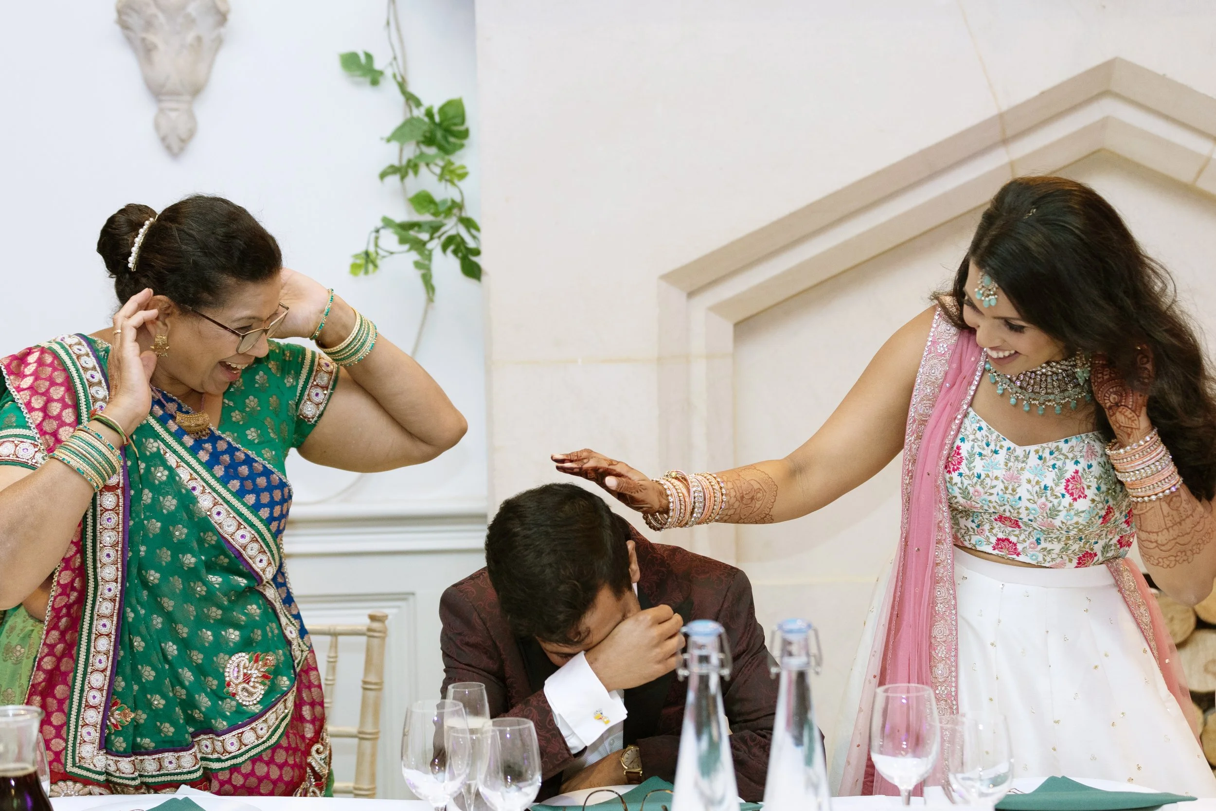 Three women and one man at a celebration table, with two women smiling and playfully touching the man's head. The man is seated, holding his head with his hand, and is surrounded by glasses and bottles on the table. The women are dressed in traditional colorful attire and jewelry, with henna on one woman’s arm.