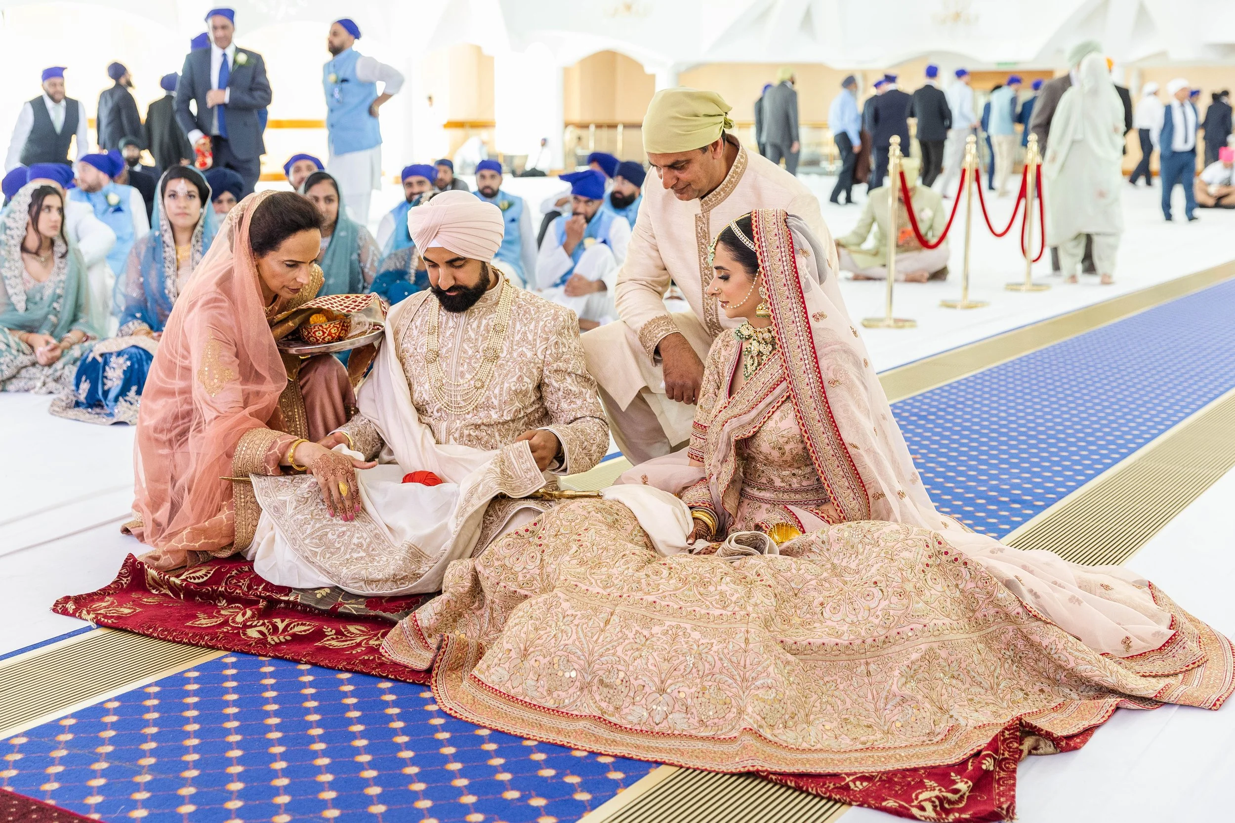 Indian wedding ceremony with bride and groom seated on the floor, dressed in traditional wedding attire, surrounded by family and friends in colorful clothing, in an ornate indoor setting.