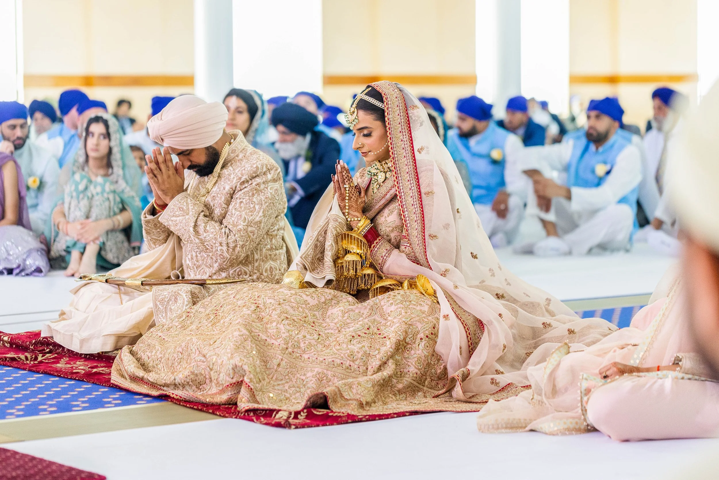 A Sikh wedding ceremony with the bride and groom kneeling on the floor with bowed heads, hands pressed together in prayer, surrounded by guests wearing traditional attire.