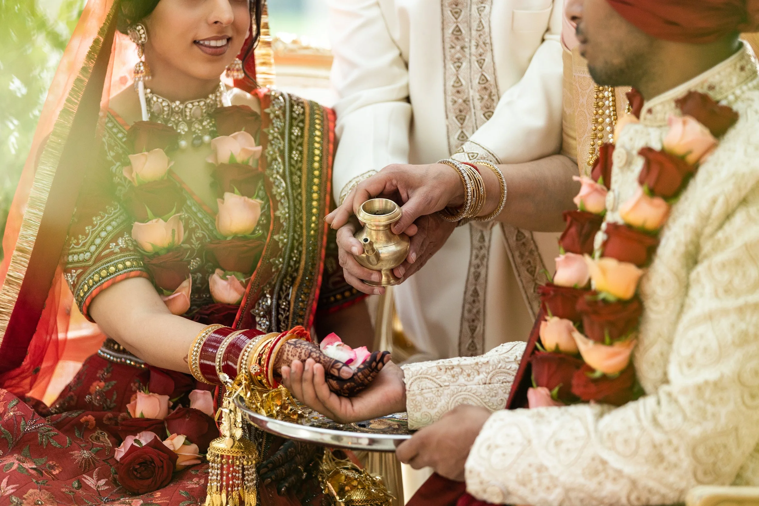 Indian bride and groom during wedding ceremony, exchanging flower garlands and holding a sacred vessel, with officiant present.