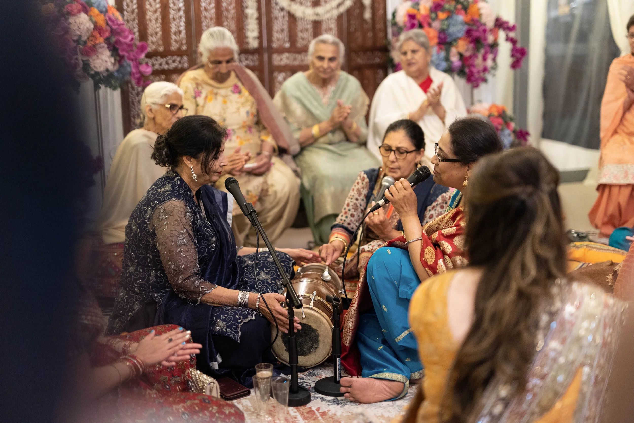 Group of women dressed in traditional Indian attire participating in a singing or prayer session, sitting on the floor with microphones and musical instruments, surrounded by floral decorations.