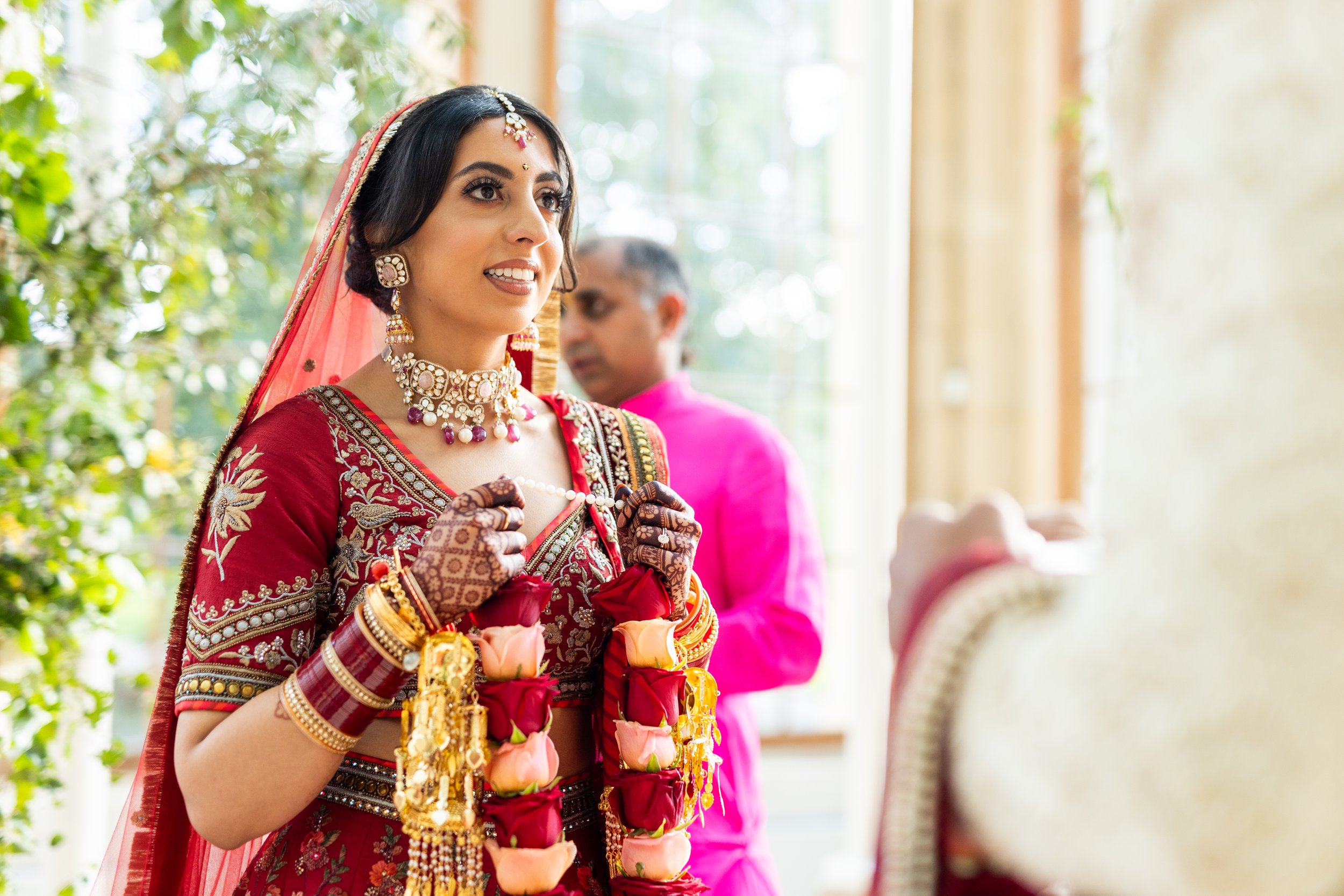 A woman dressed in traditional Indian wedding attire, wearing jewelry and mehndi on her hands, holding a garland of roses, standing indoors during a wedding ceremony.