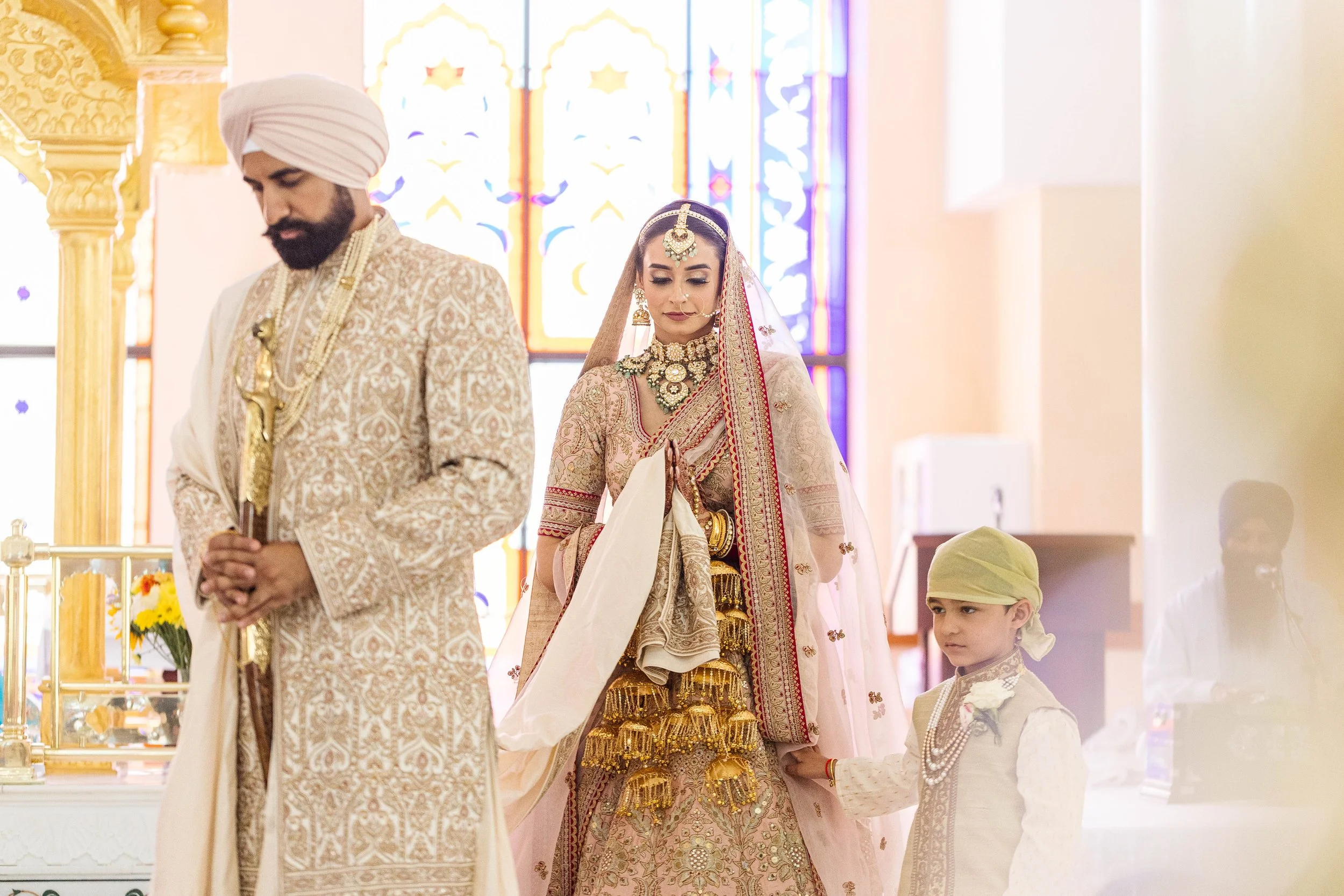 A traditional Indian wedding ceremony with a bride, groom, and young boy in ceremonial attire inside a decorated temple, with stained glass windows in the background.