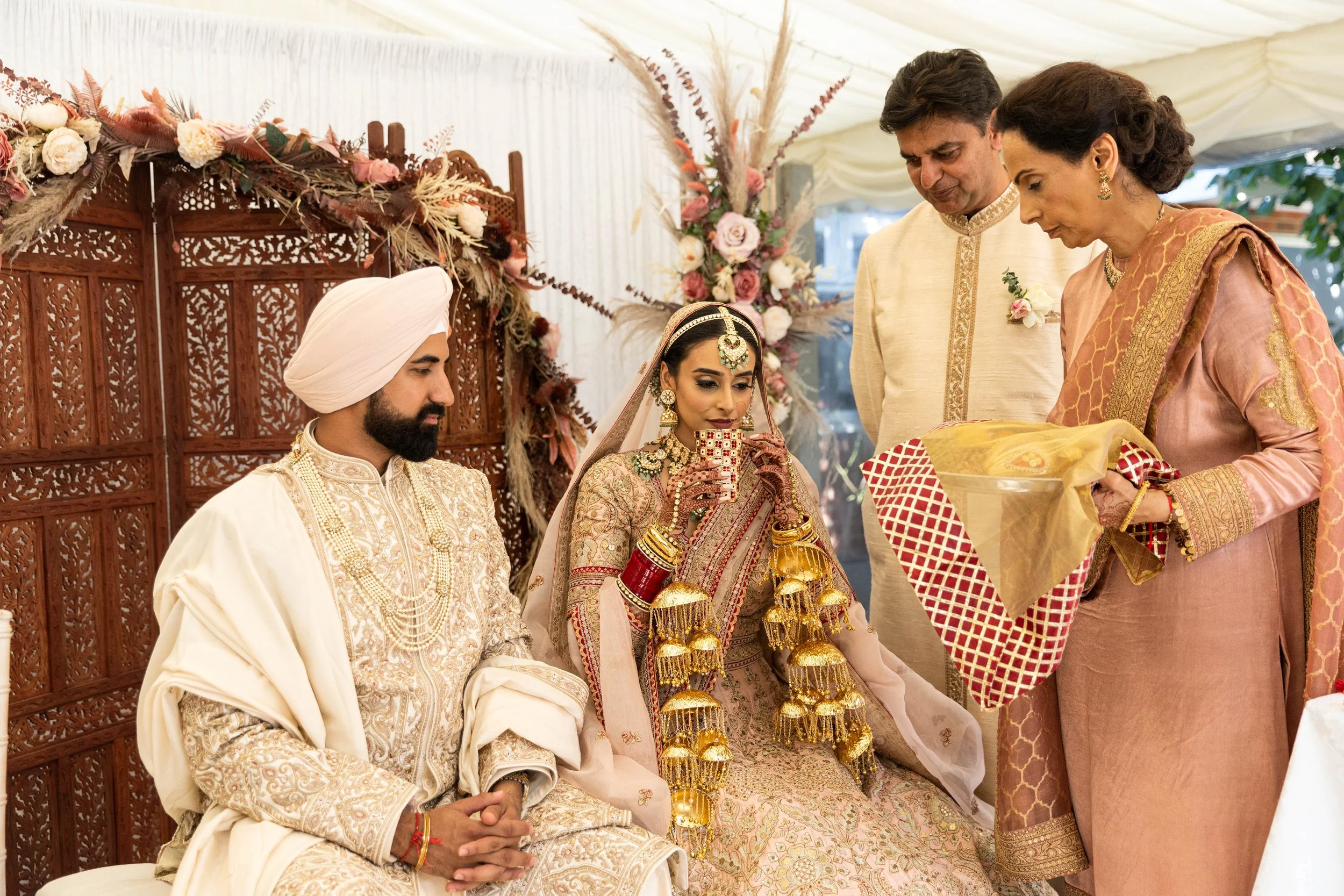 Indian wedding ceremony with bride and groom dressed in traditional attire, seated on a decorated stage, surrounded by family members, during a gift exchange.