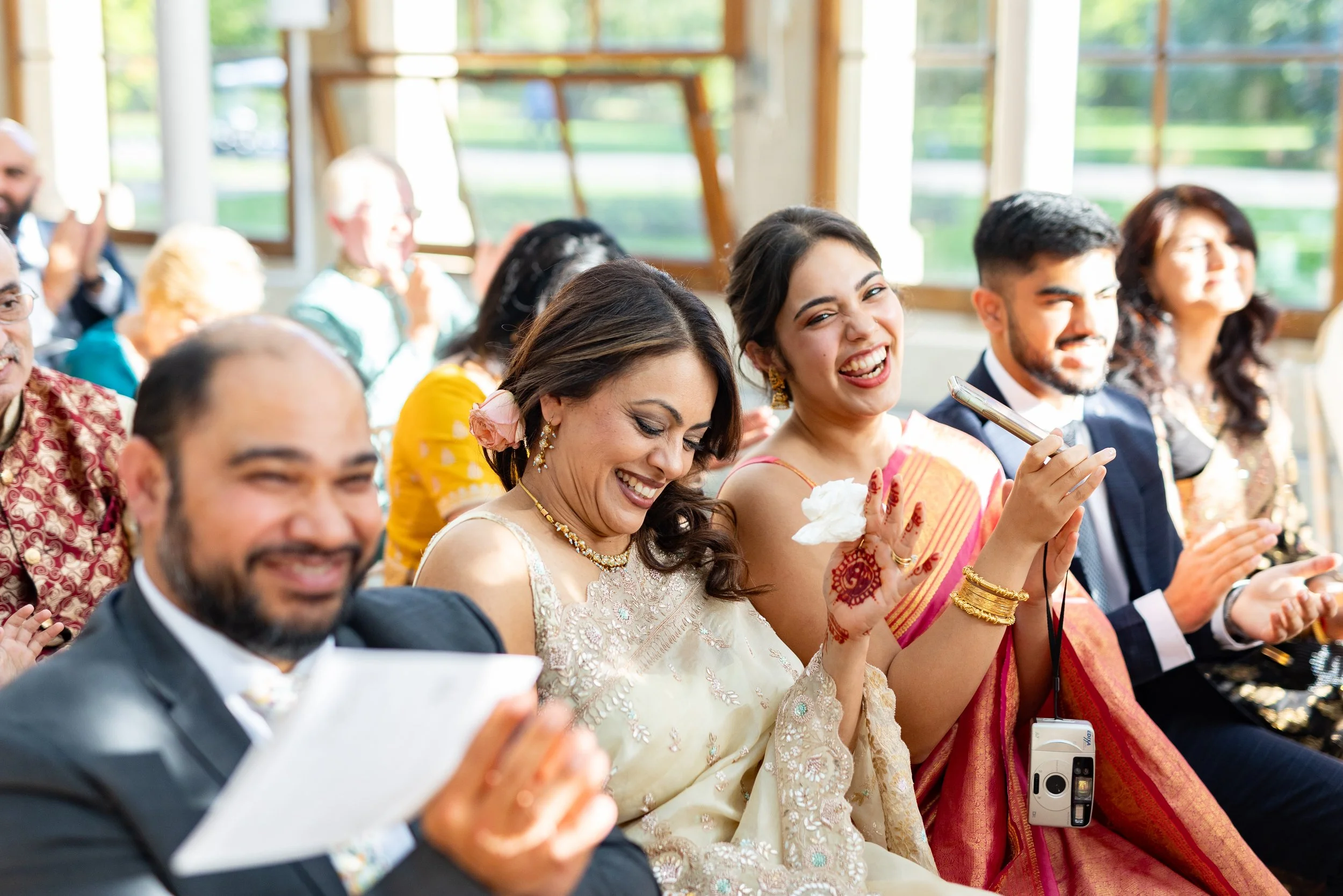 People dressed in traditional Indian attire attending an event, laughing, and clapping indoors with windows showing greenery outside.