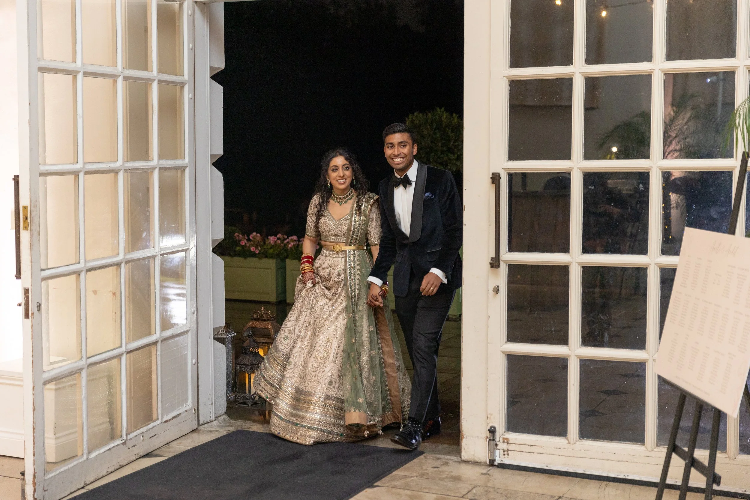 A couple dressed in formal wedding attire standing together at the entrance of a building at night. The woman is wearing a traditional gold and green Indian dress with jewelry and henna, and the man is wearing a black tuxedo with a bow tie.