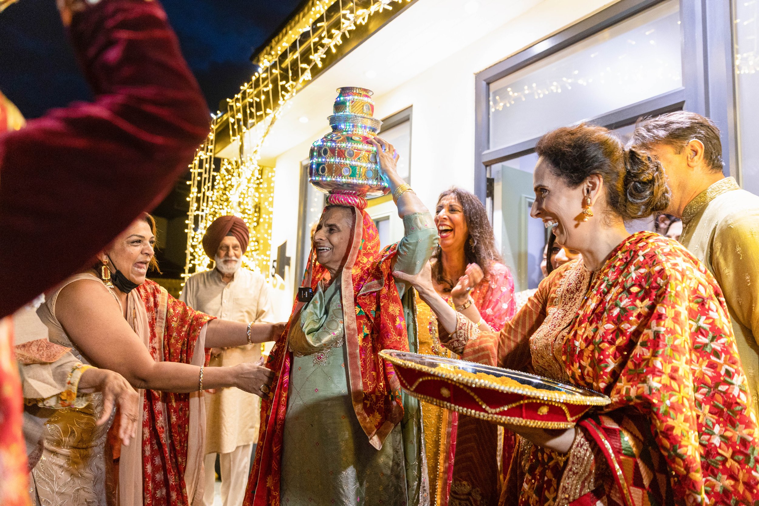 Group of people celebrating an Indian birthday party, with an elderly woman holding a decorated silver pot on her head, surrounded by smiling family and friends.