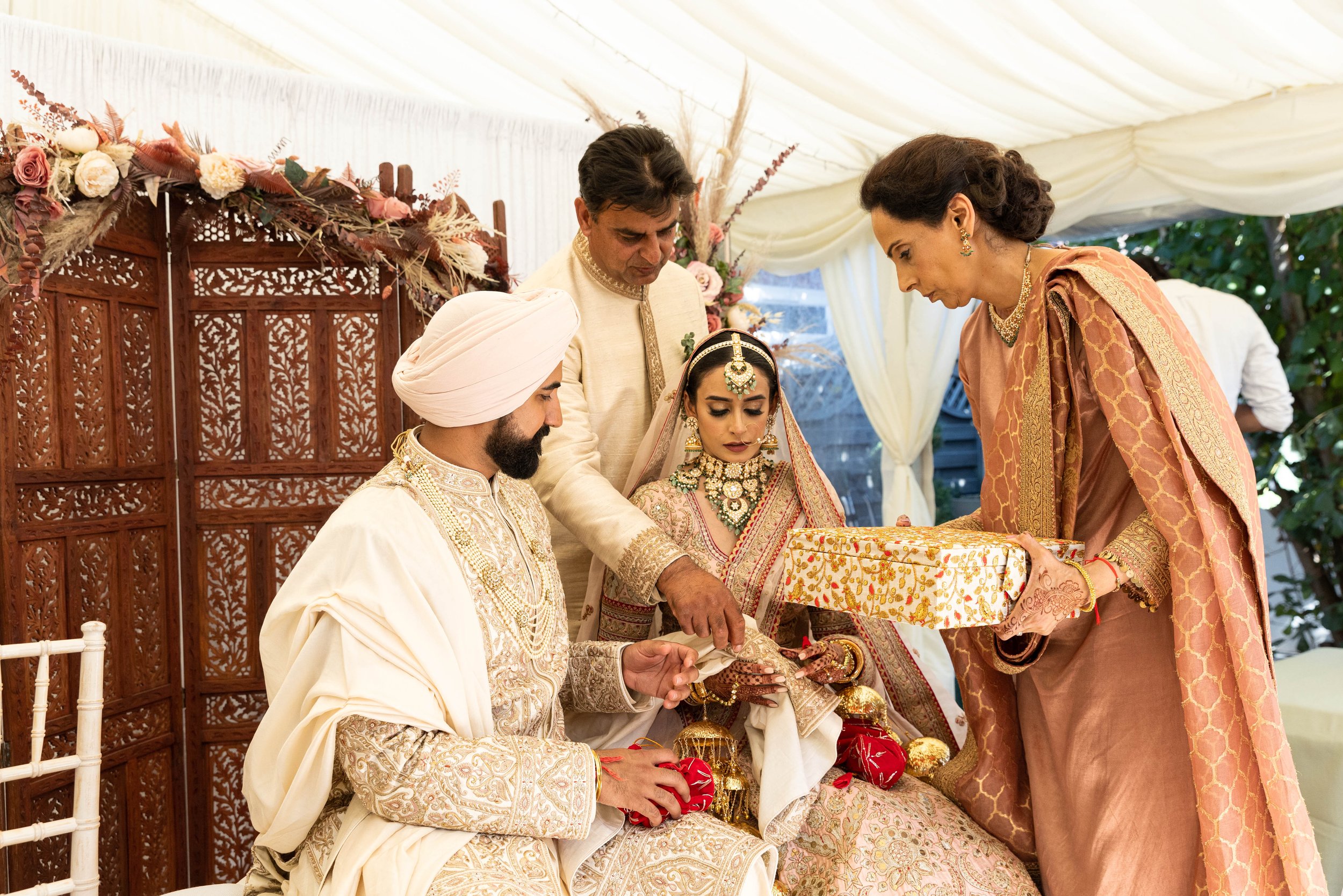 Indian wedding ceremony with bride and groom in traditional attire, surrounded by family members exchanging gifts under a decorated tent.