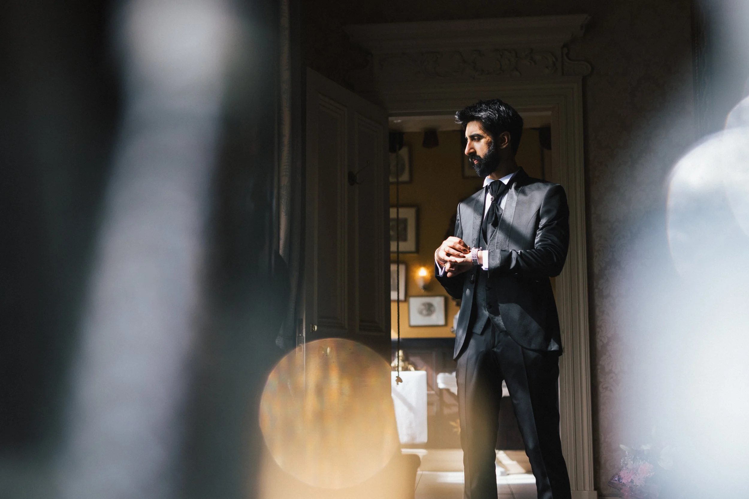 A man with a beard wearing a formal black tuxedo, standing inside a room, adjusting his cufflinks, with framed pictures on the wall behind him.
