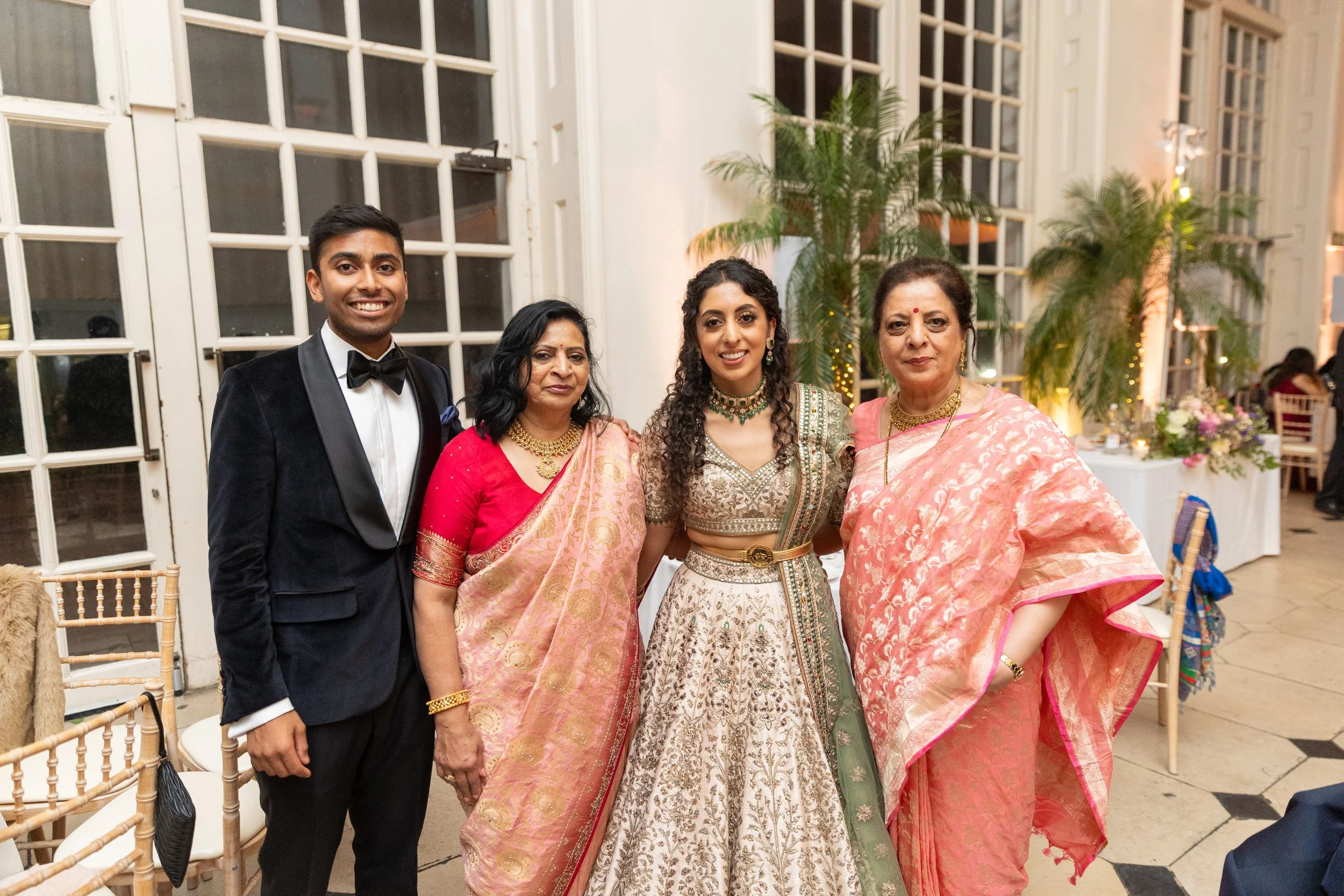 Five people standing together in formal attire at a celebration, with large windows, plants, and decorated tables in the background.