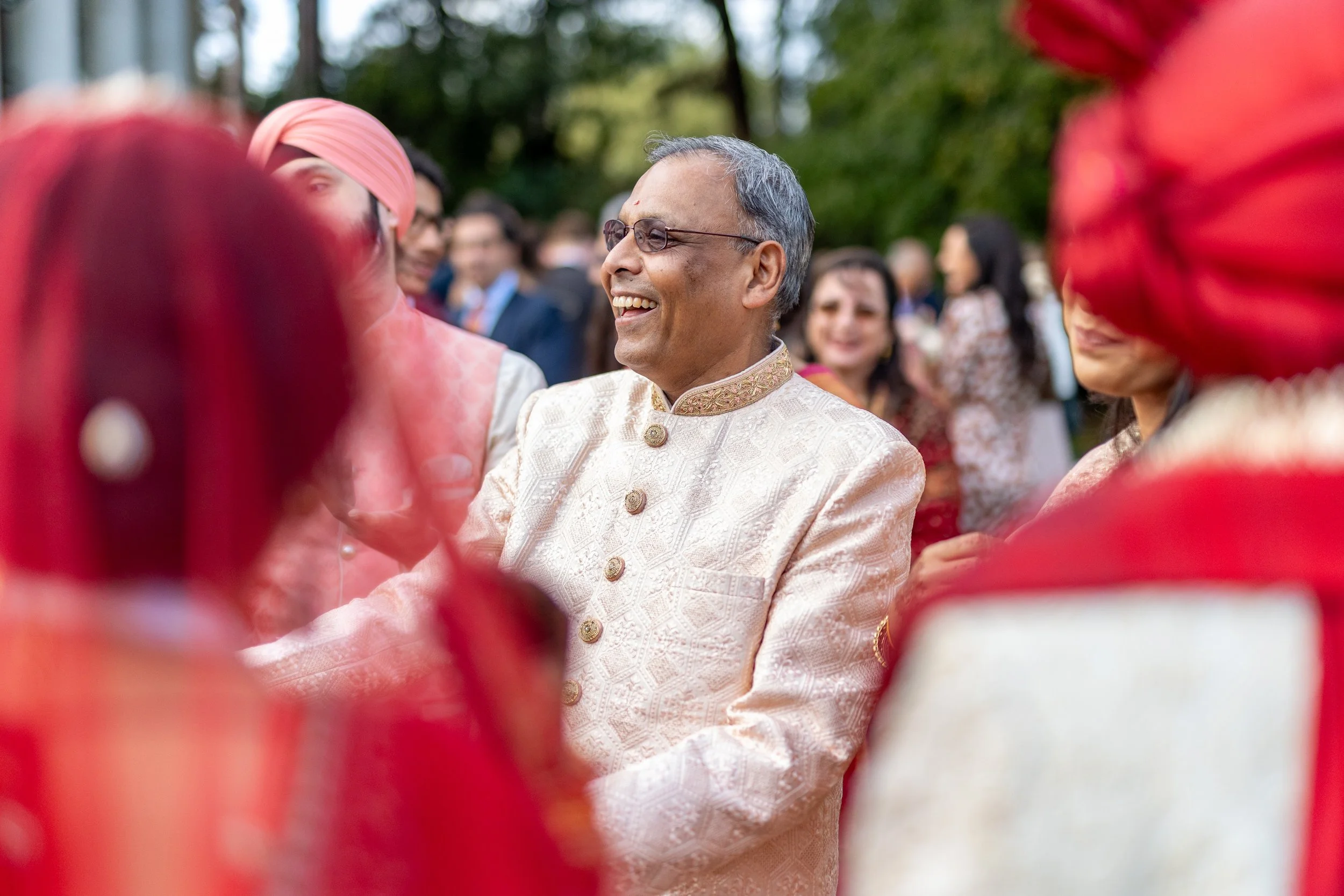 A man in traditional Indian attire smiling and greeting at an outdoor celebration or wedding, surrounded by women and men in colorful traditional clothing.