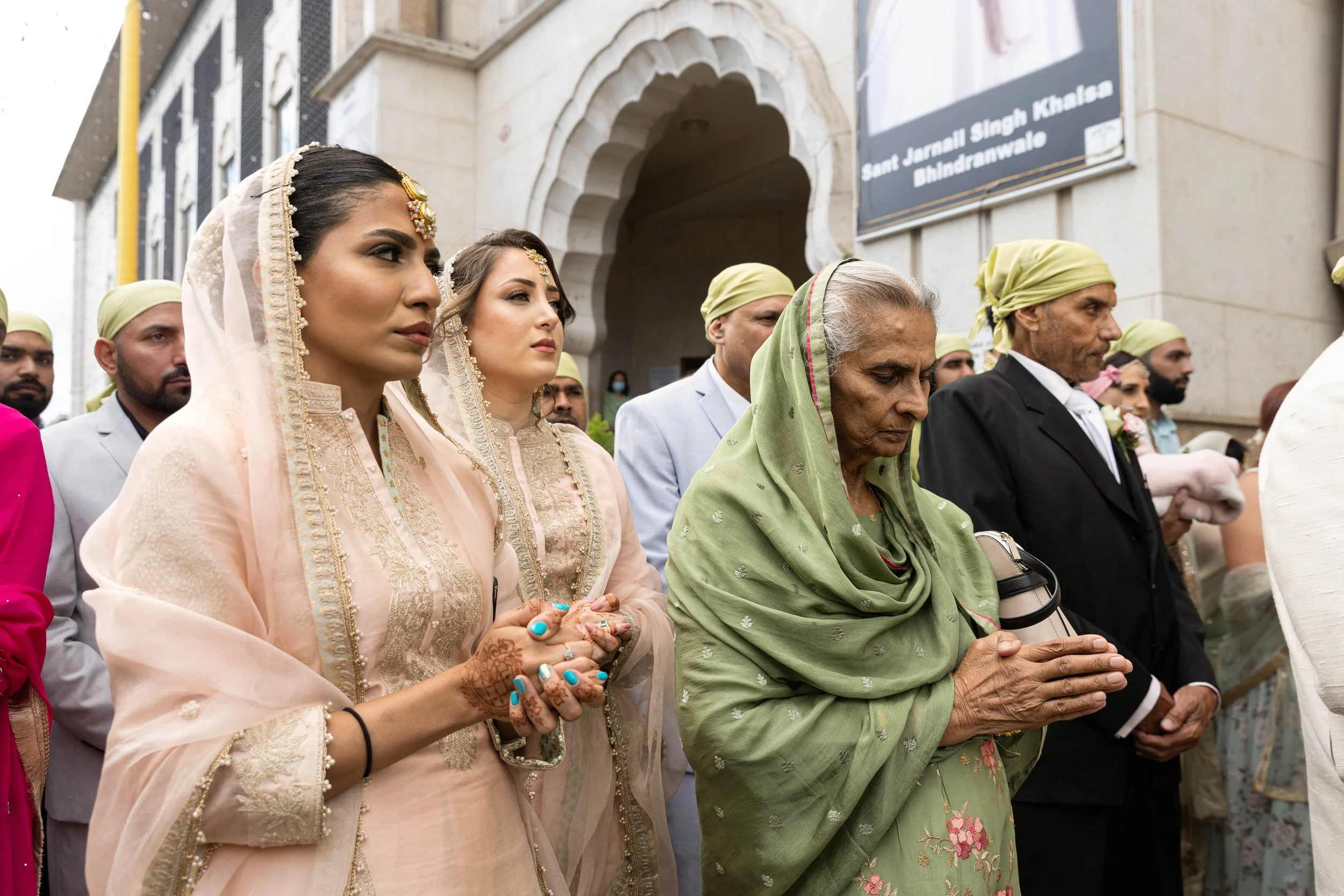 People participating in a religious or cultural ceremony outside a building, with women in traditional Indian attire and men in formal suits, some with head coverings.