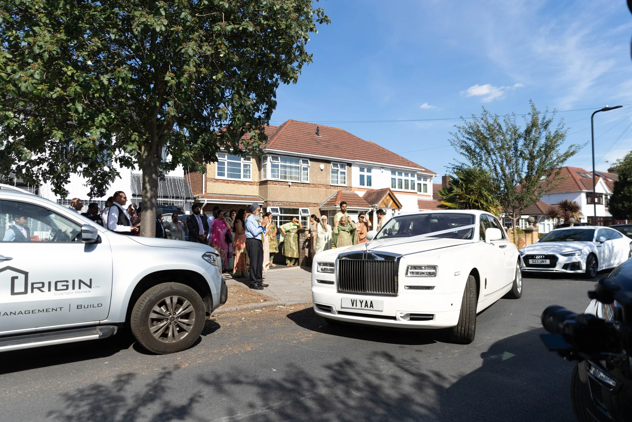 A white luxury car with a custom license plate 'VI YAA' parked on a residential street during daytime, with a crowd of people dressed in formal and traditional attire gathered in the background outside a row of houses, and several other cars parked a