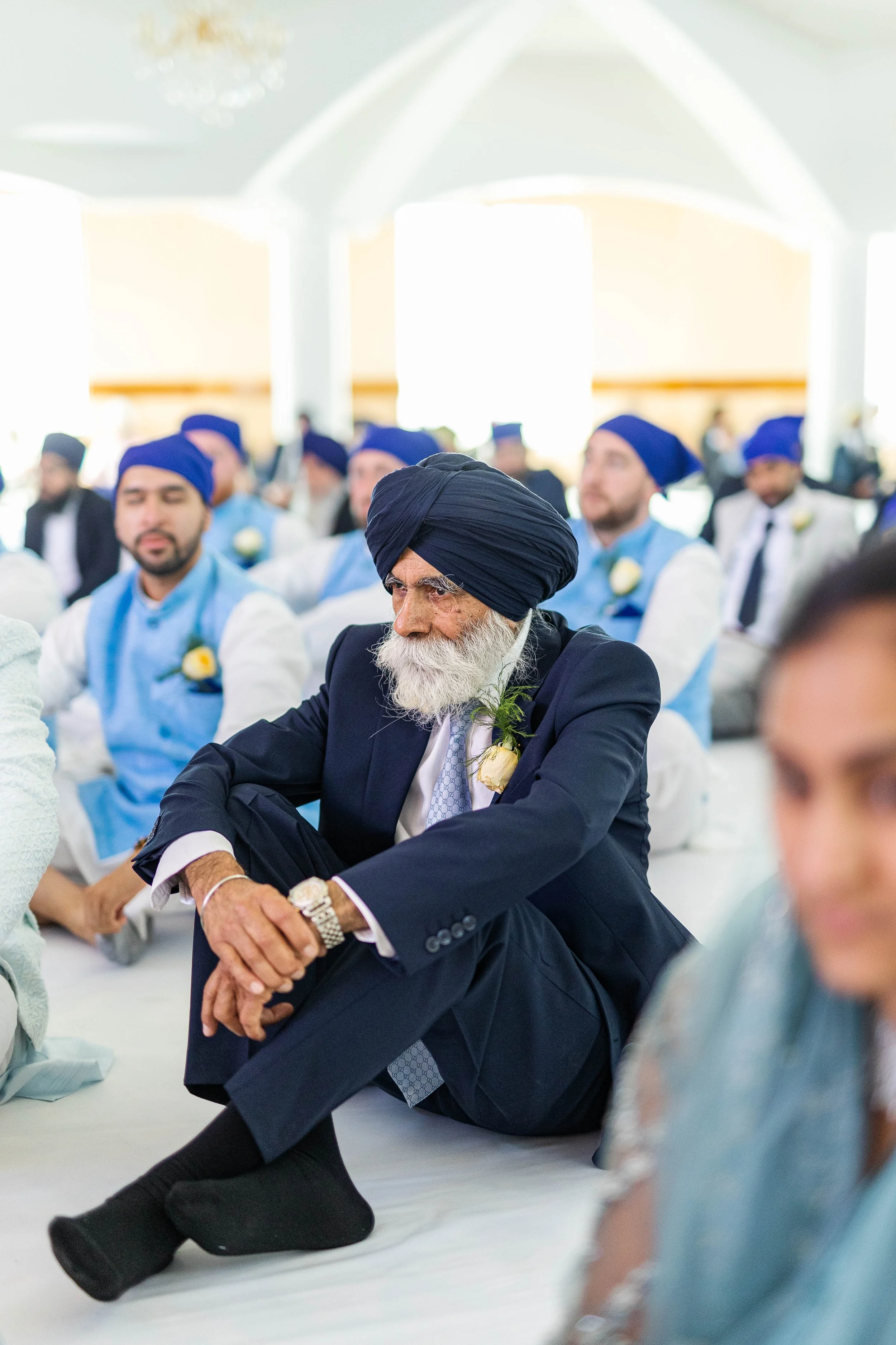An elderly Sikh man with a white beard, dressed in a dark suit and a blue turban, sitting with his legs crossed during a cultural or religious ceremony, surrounded by other men in traditional Blue turbans and light-colored clothing.