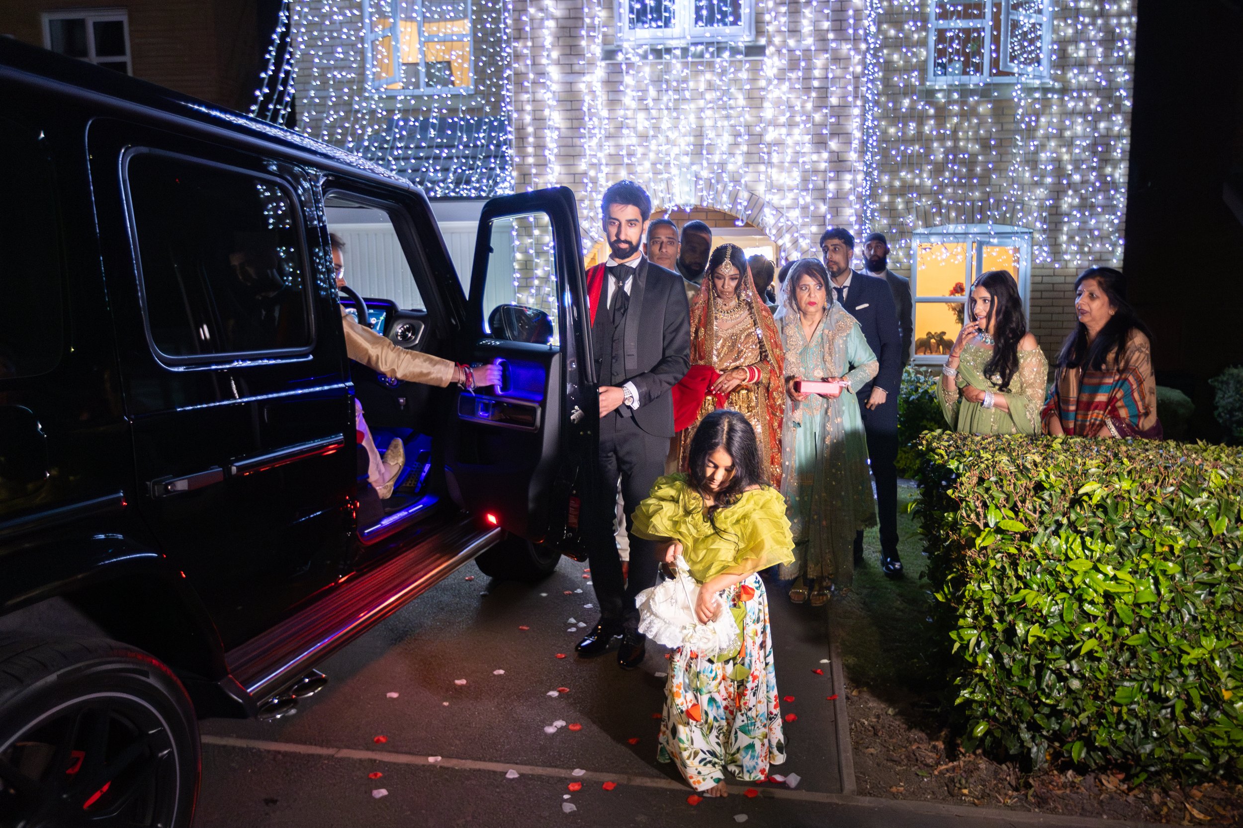 People dressed in traditional and formal attire arriving at a celebration outside a house decorated with white string lights, with a black vehicle parked nearby.