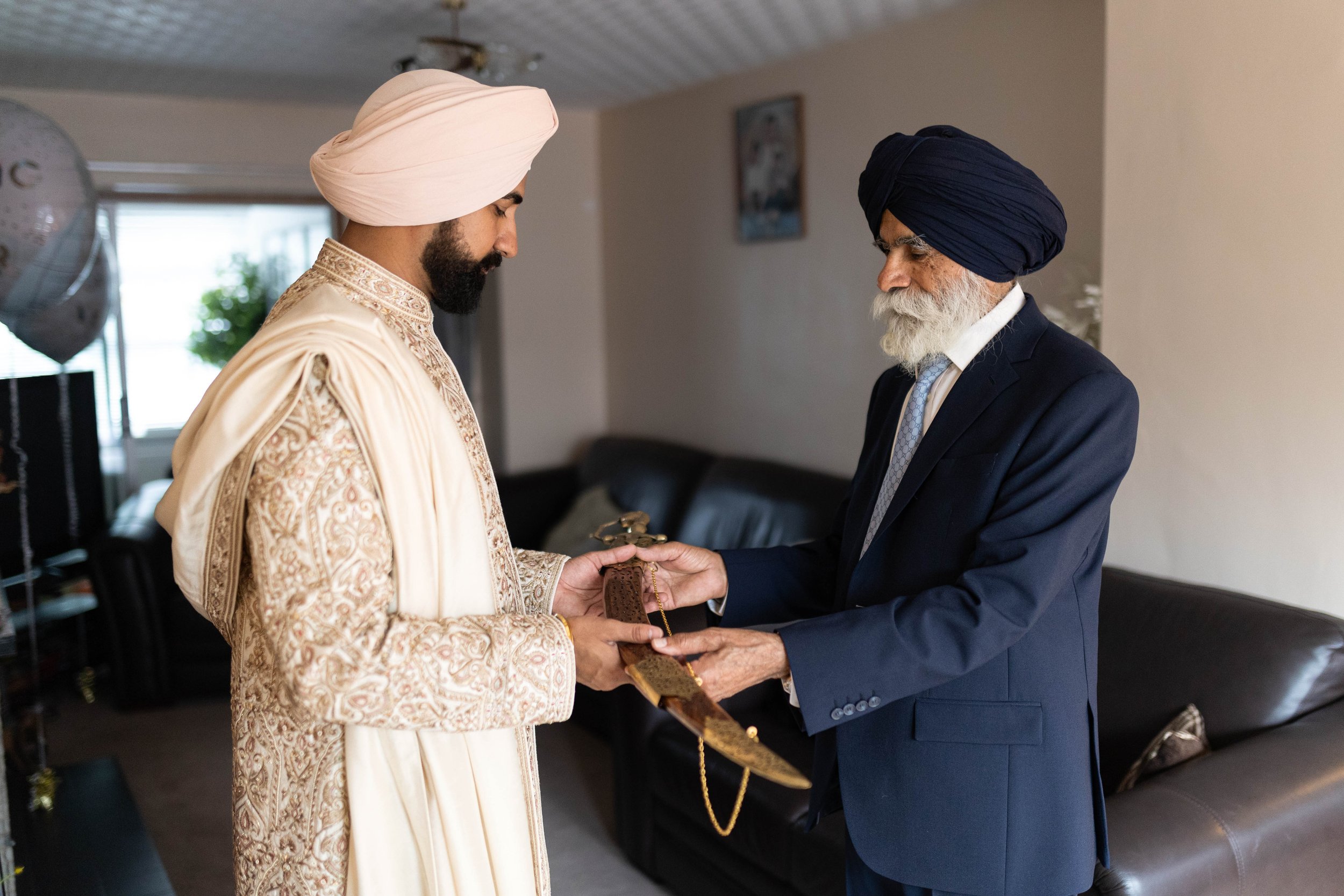 Two men dressed in traditional and formal attire engaged in a ceremonial exchange of a sword in a living room.
