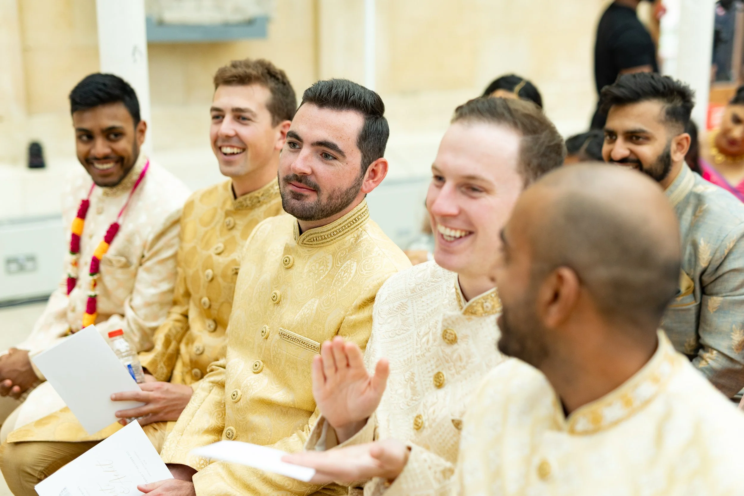 Group of men dressed in traditional Indian attire, sitting together in a celebration or ceremony, smiling and engaging with each other.