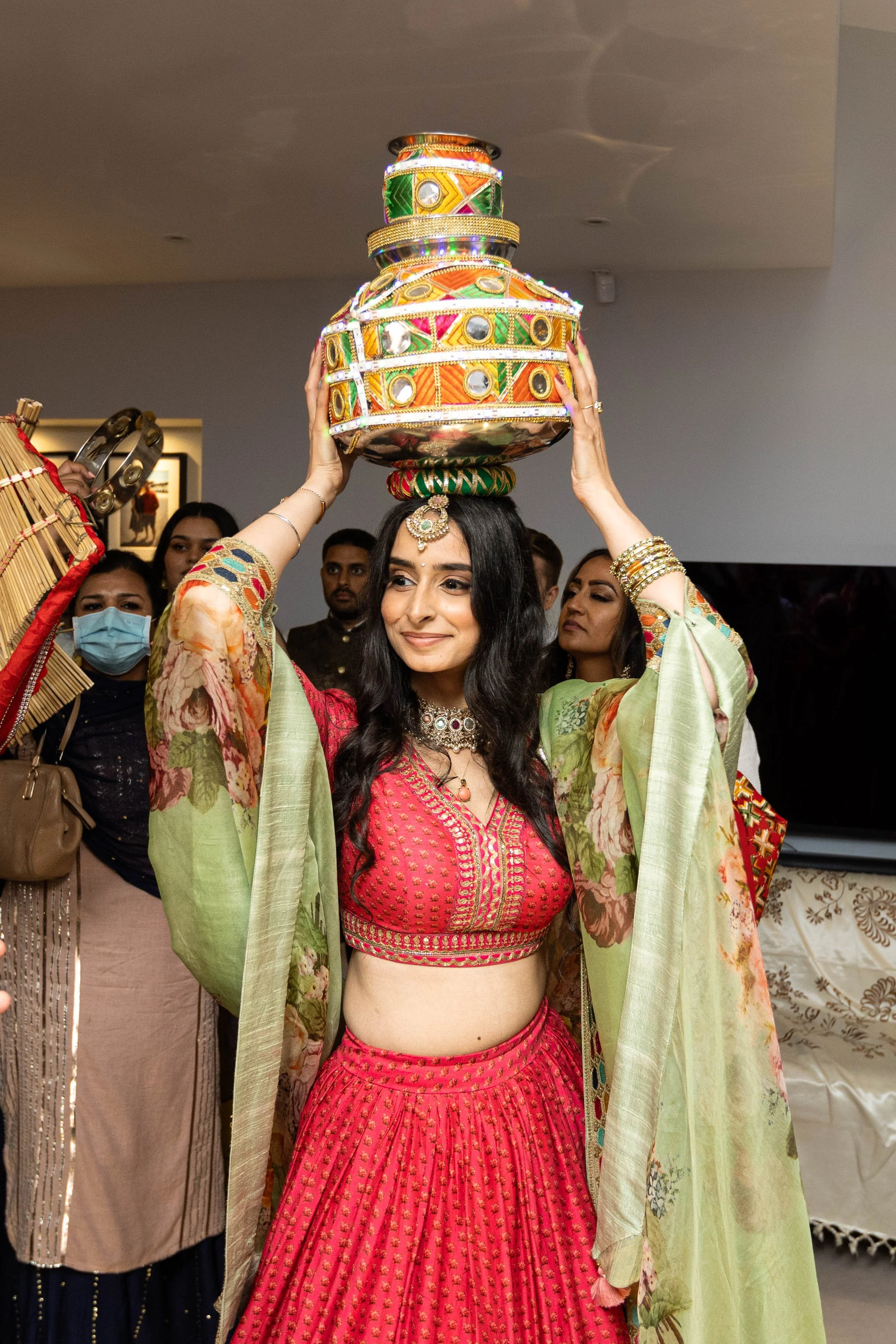 A woman dressed in traditional Indian attire, a pink and gold lehenga, is balancing a colorful decorative pot on her head during a celebration. She is surrounded by people, some of whom are wearing masks.