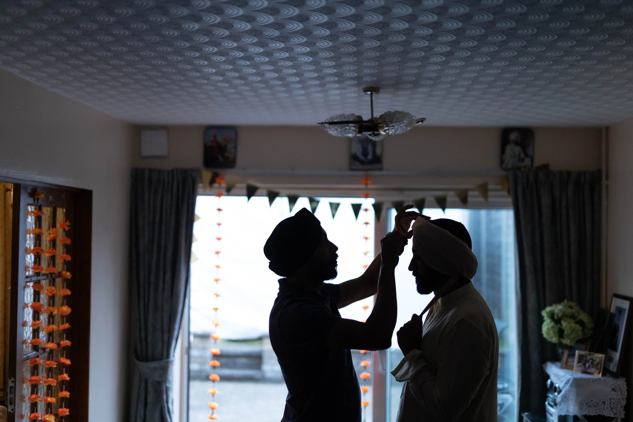 Silhouette of a person tying a turban on another person's head inside a home, with decorative curtains and photos in the background.