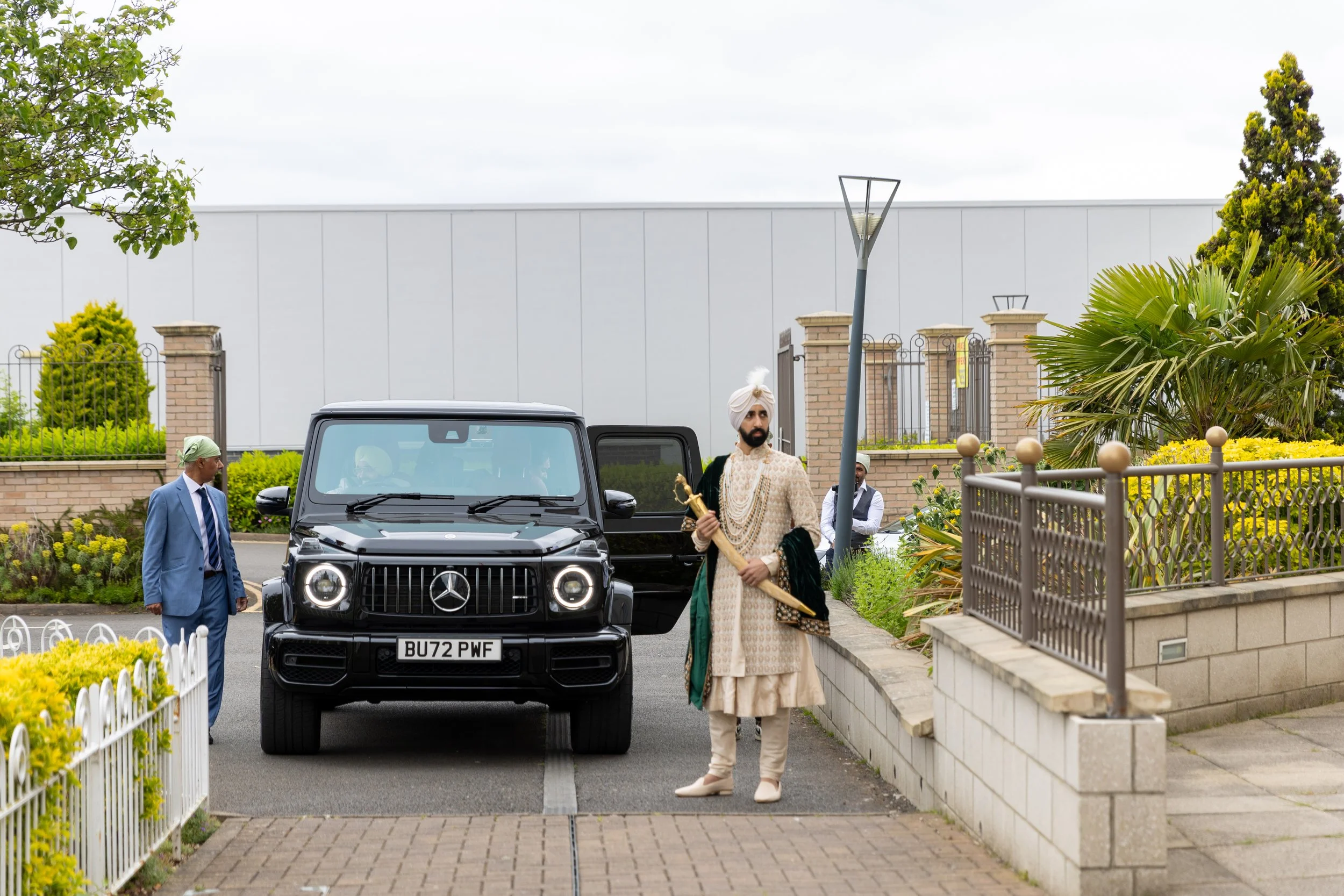 Man dressed in traditional Indian attire holding a sword, standing beside a black Mercedes-Benz G-Class vehicle with the license plate 'BU72 PWF', in a landscaped area with yellow and green plants, brick walls, and lampposts.