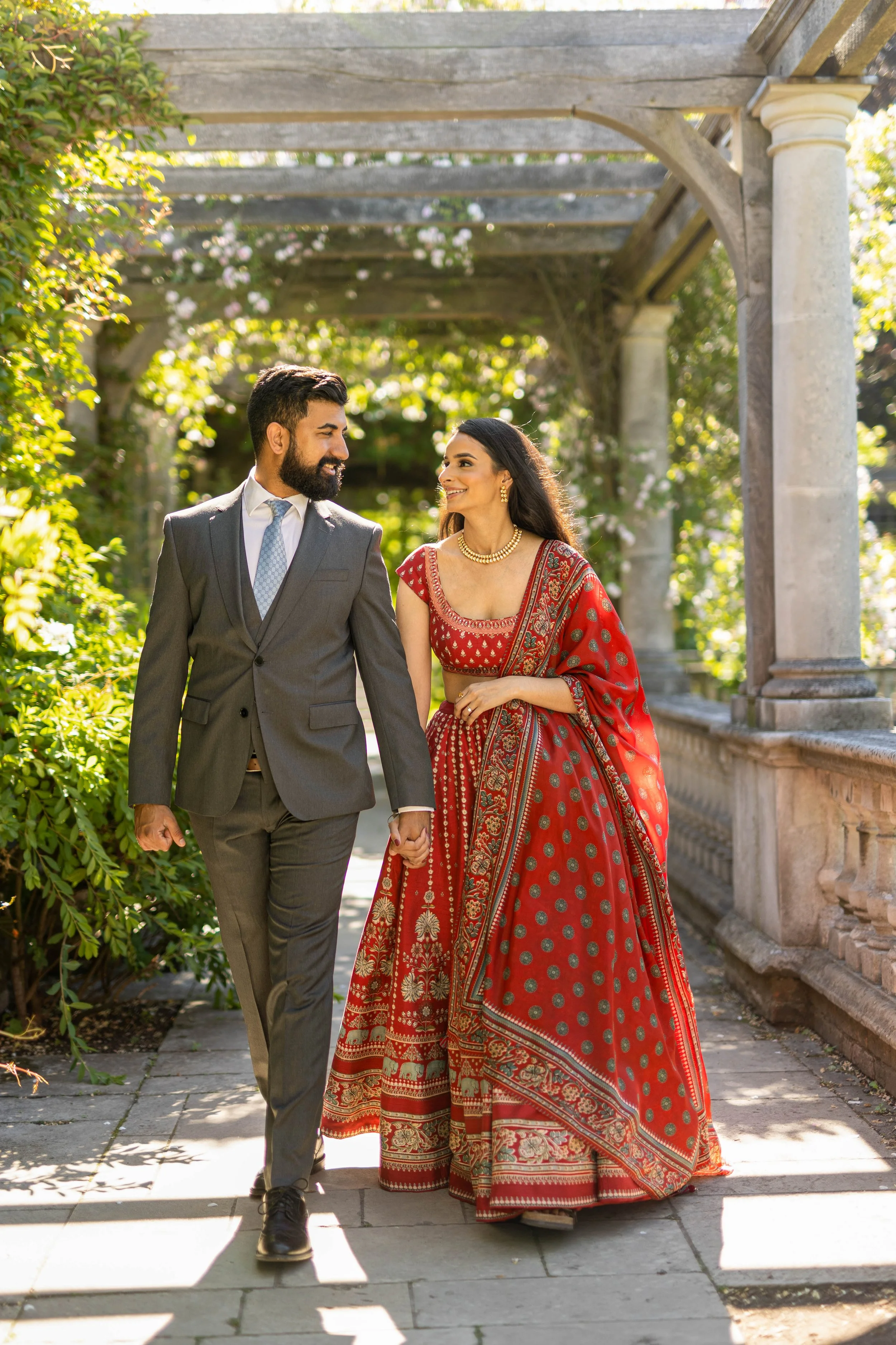 A couple walking hand in hand outdoors in a garden with a wooden pergola in the background. The woman is wearing a traditional red Indian saree with gold accents, and the man is dressed in a gray suit with a tie.