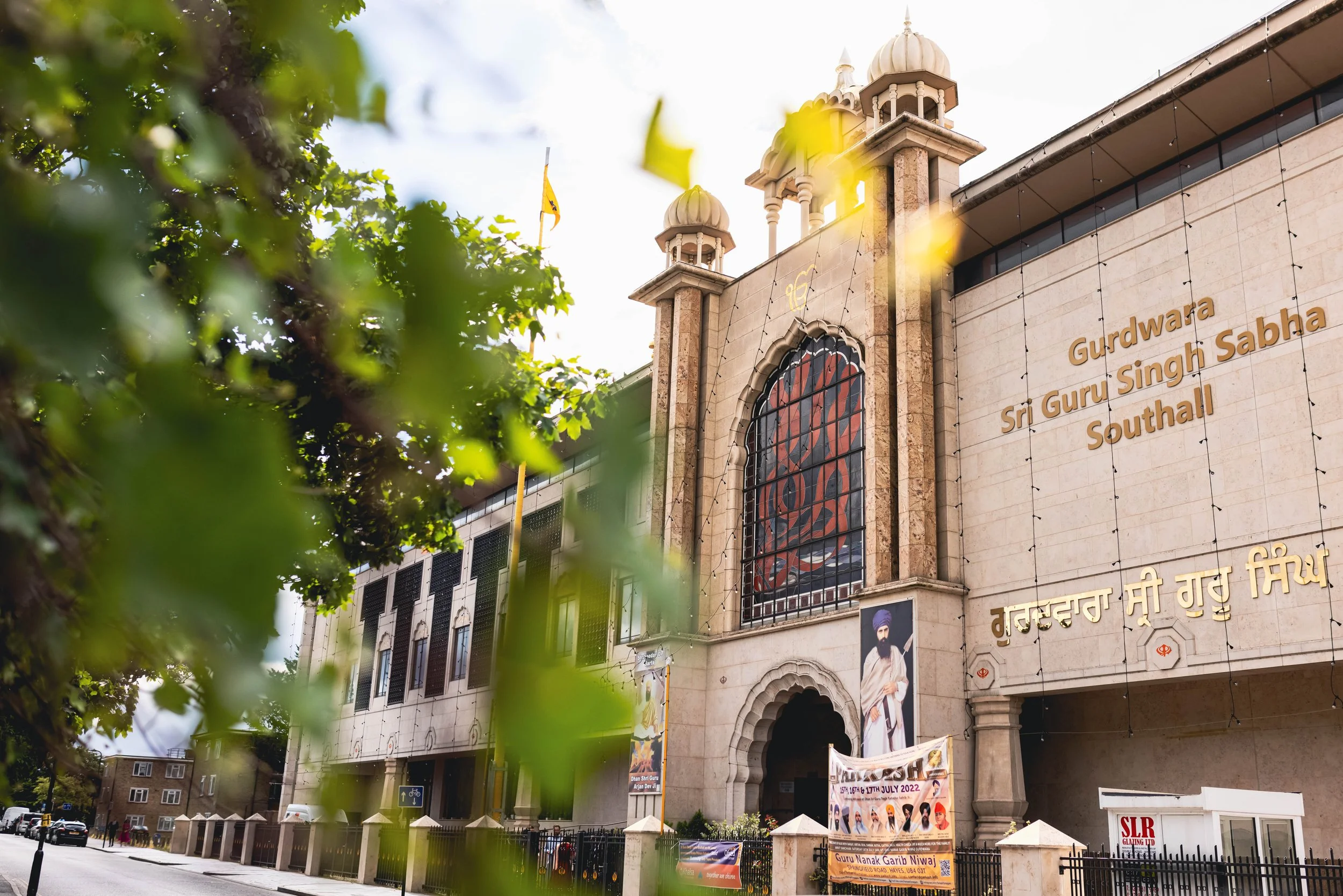 The exterior of the Gurdwara Sri Guru Singh Sabha Southall, a Sikh temple, with large gold lettering on the building's wall, a picture of a Sikh spiritual leader near the entrance, and a large stained glass window. The scene is partly obscured by gre