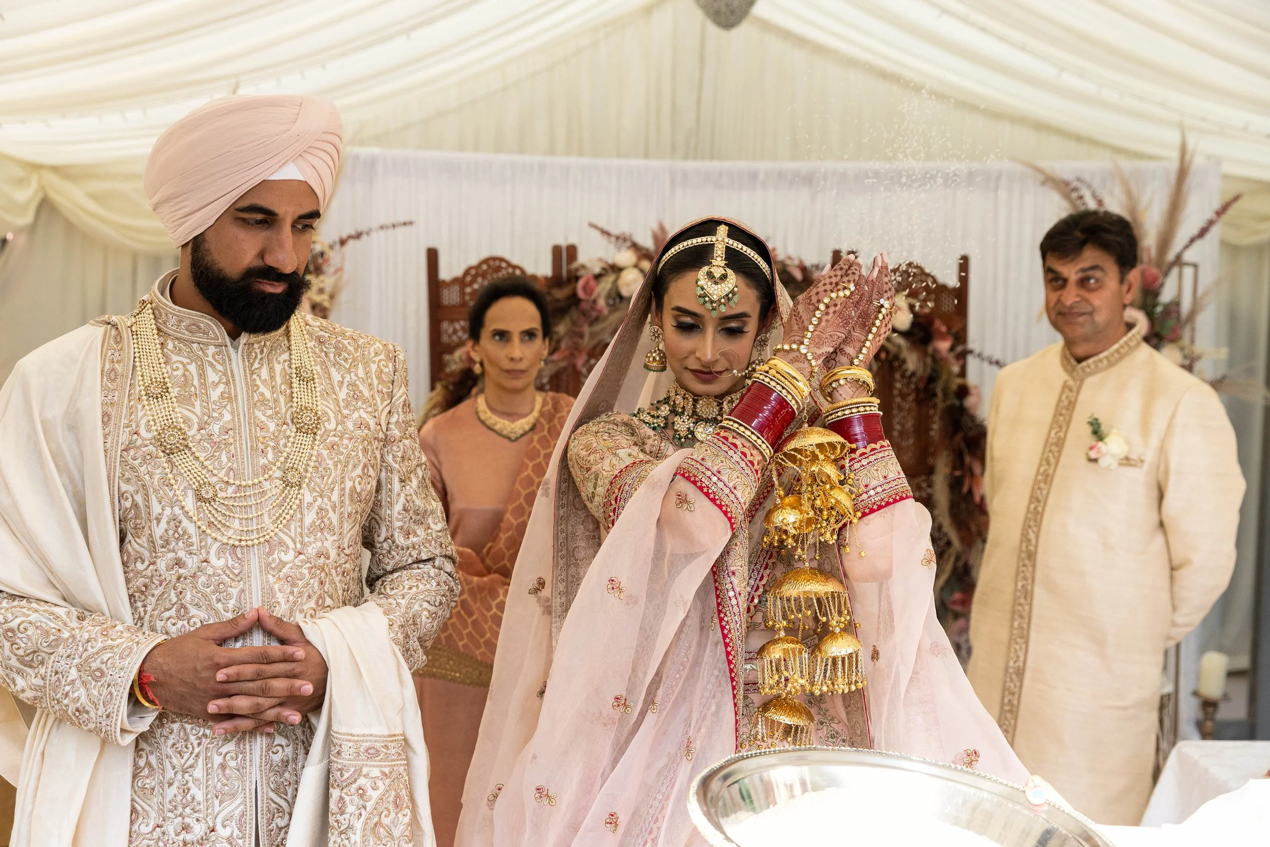 An Indian bride performing a traditional ritual at her wedding, surrounded by family members, inside a decorated tent.