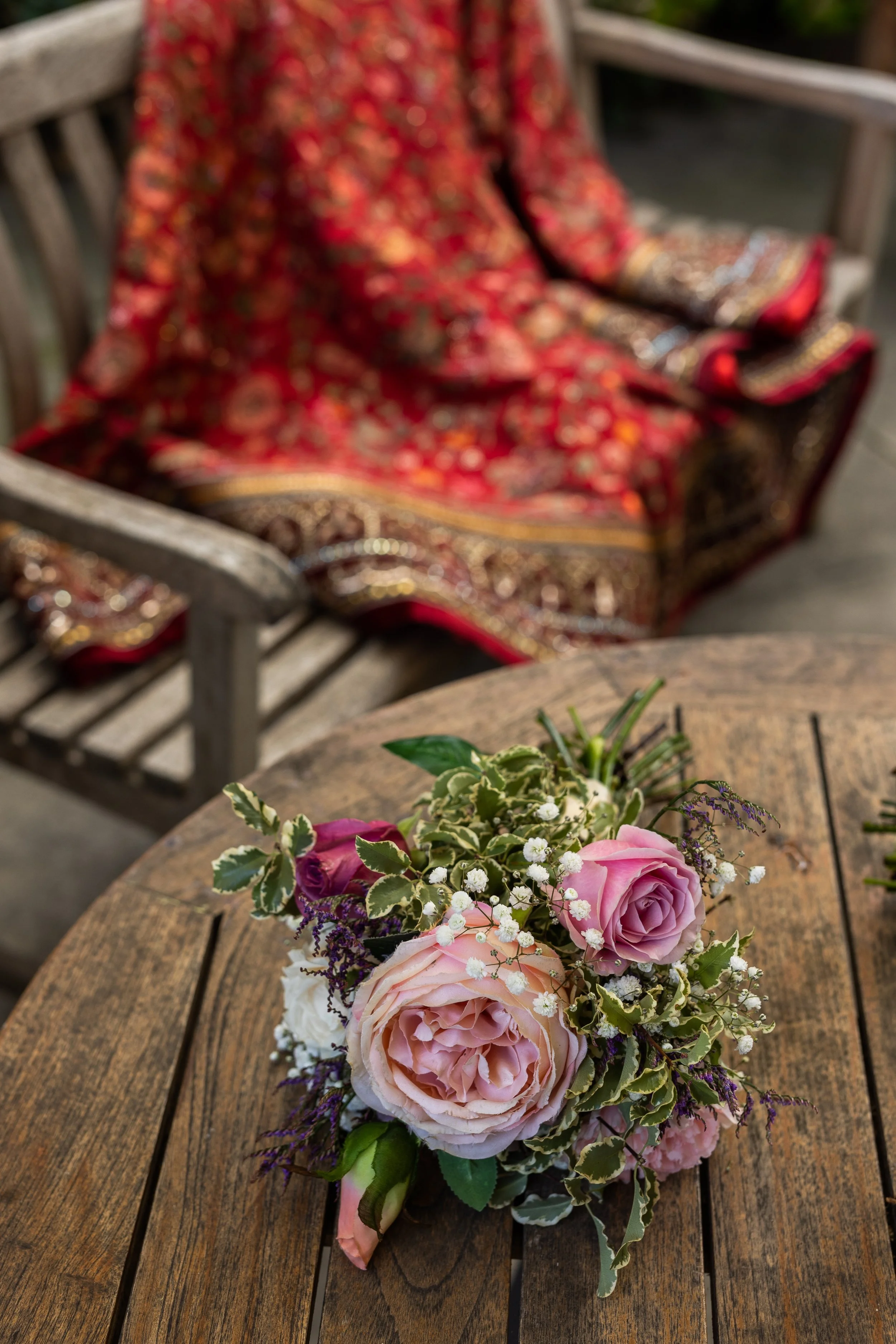 A bouquet of pink and purple roses with greenery and small white flowers on a wooden table, with an ornate red patterned fabric and wooden chair in the blurred background.
