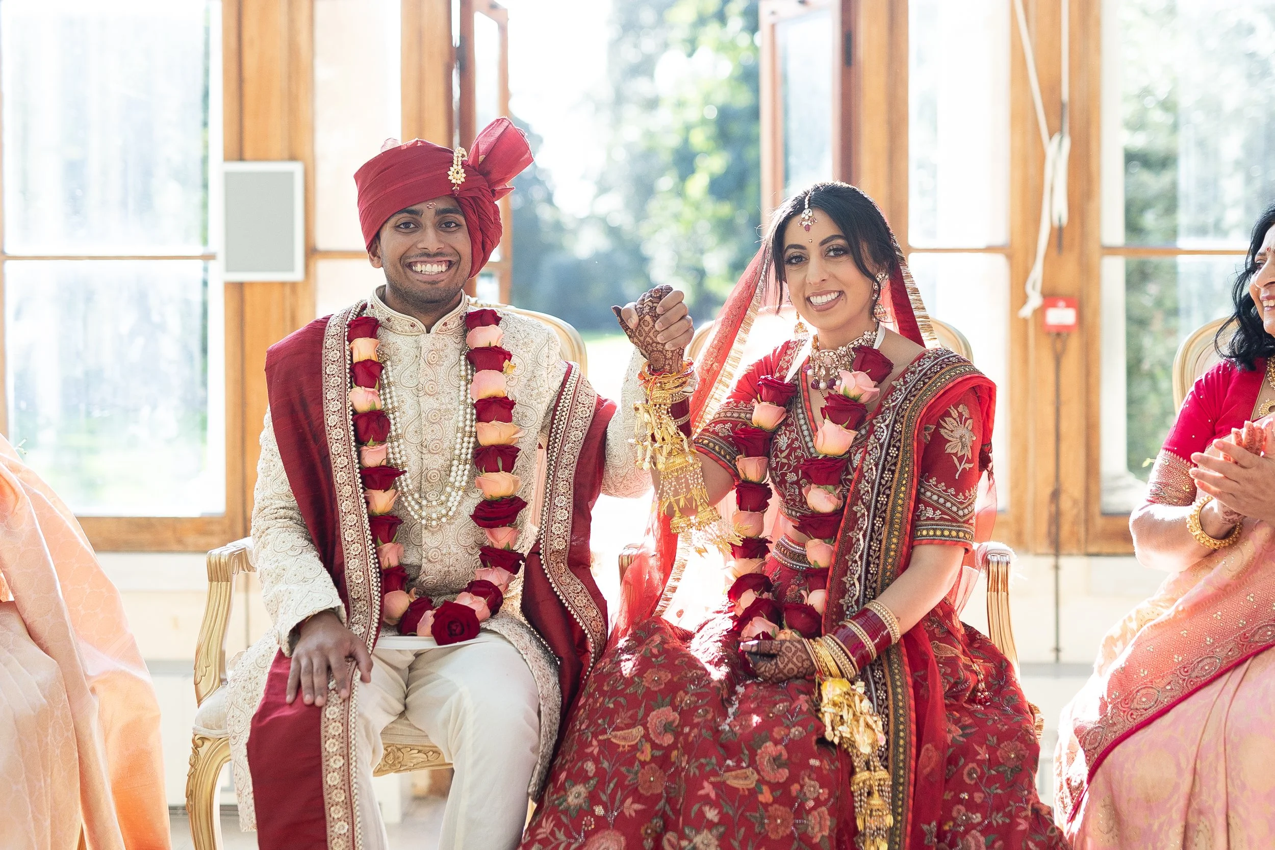 A newlywed Indian couple sitting on chairs, smiling, holding hands, wearing traditional wedding attire with floral garlands, inside a wooden-paneled room with large windows and sunlight.