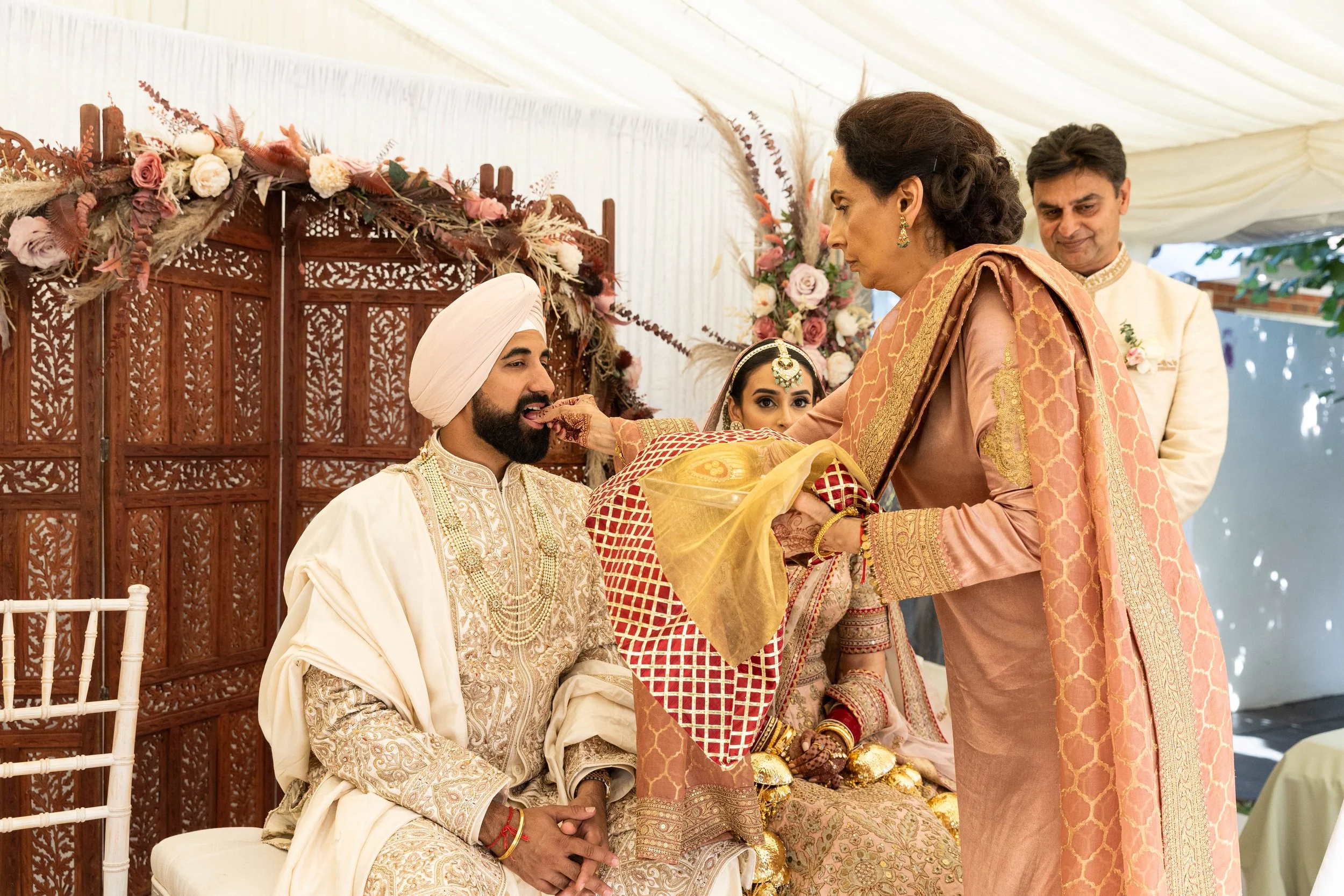 Indian groom in traditional wedding attire seated as bride seated next to him is being blessed by an older woman in an orange and gold saree during a wedding ceremony, with a man standing behind them in the background.