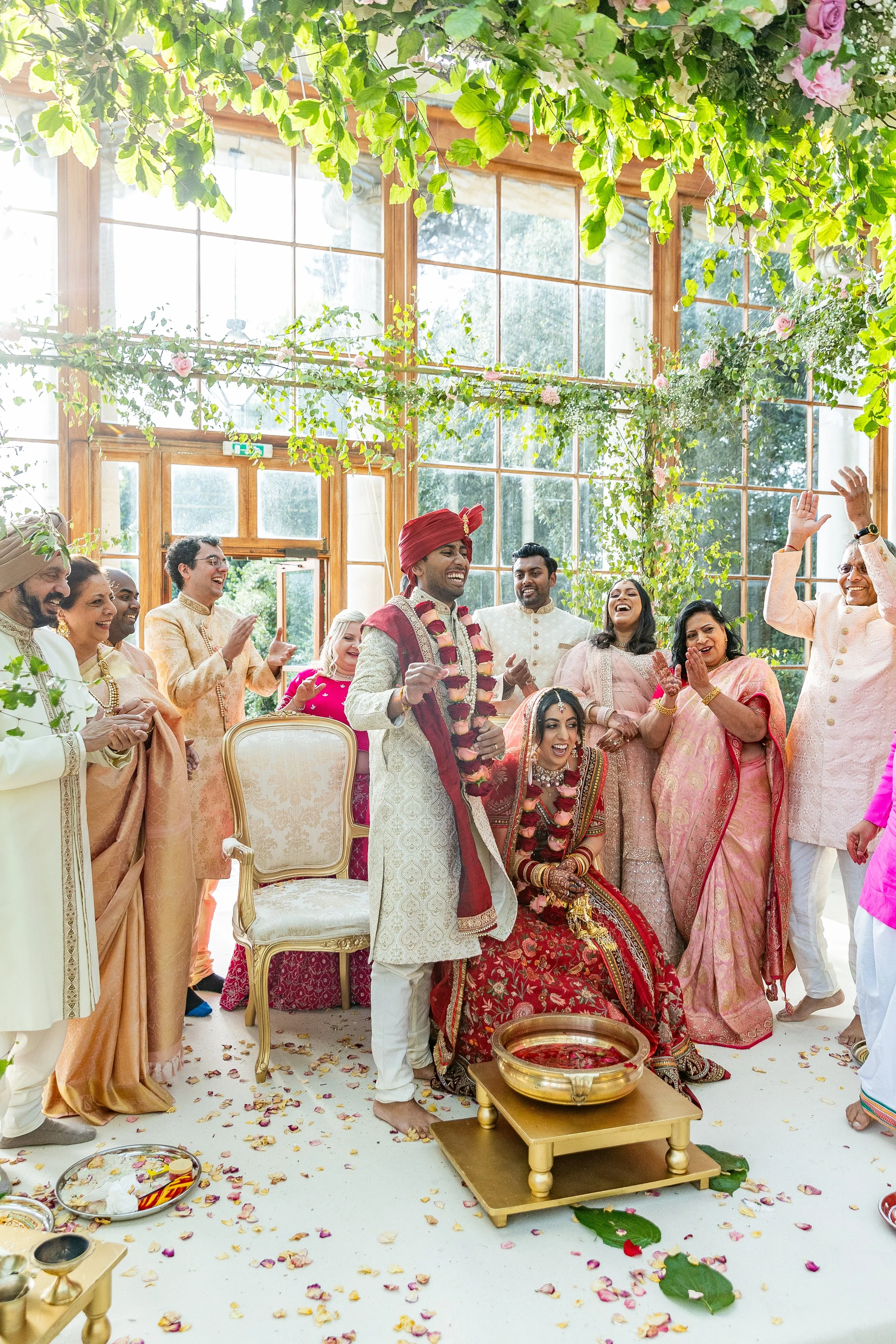 Indian wedding ceremony inside a bright, glass-ceilinged room decorated with flowers. The bride and groom are seated, dressed in traditional wedding attire, surrounded by joyful family and friends.